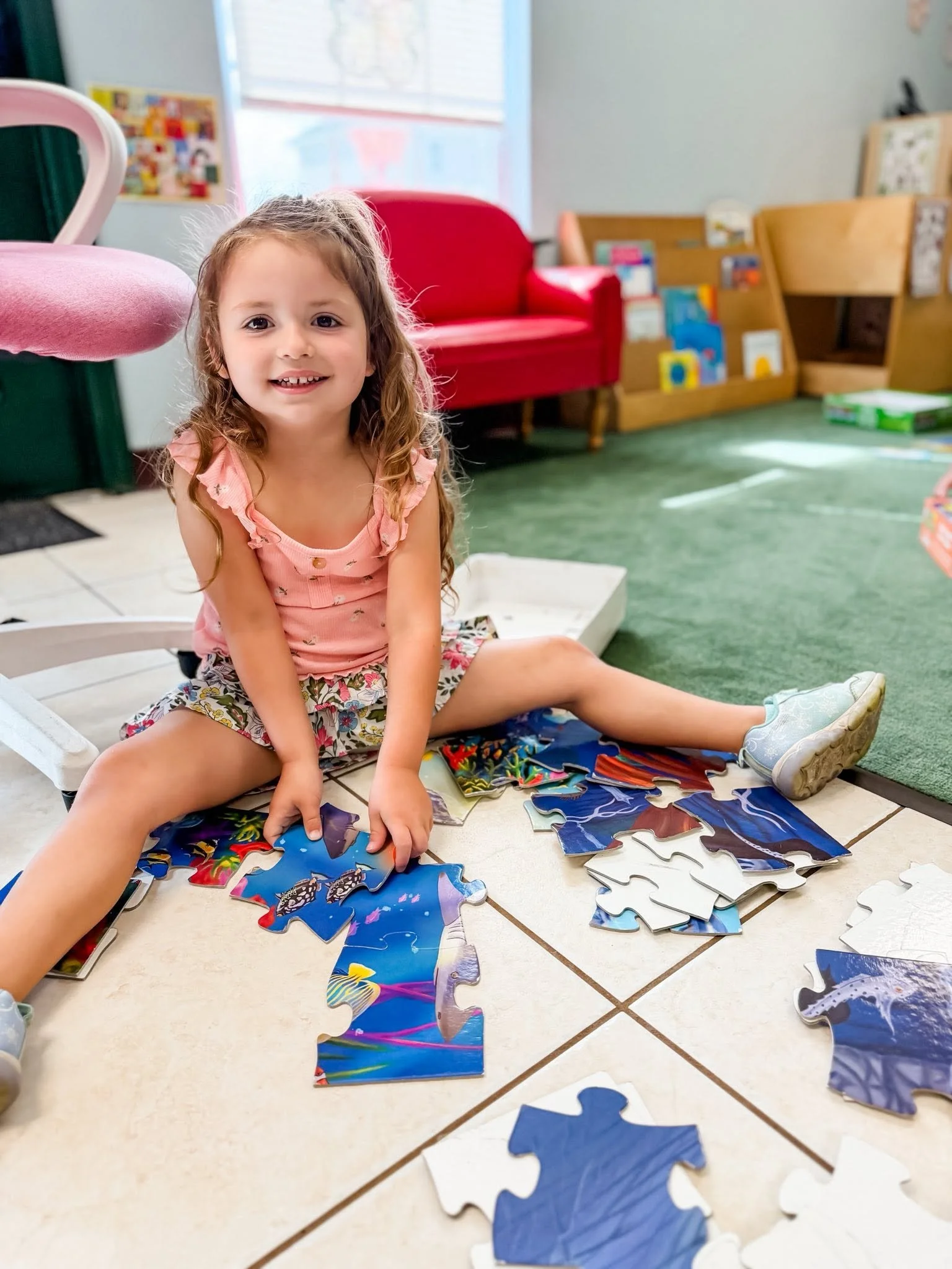 A young girl with curly hair in a pink shirt and floral shorts sitting on a tiled floor, assembling a colorful jigsaw puzzle with ocean and fish designs. Behind her, there’s a red couch, wooden bookshelves filled with books, and a window letting in natural light.