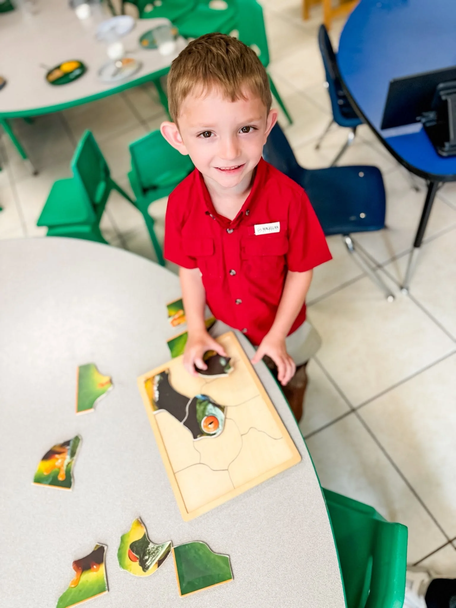 A young boy wearing a red shirt and a name tag standing at a table with a partially completed puzzle. The background includes several green and blue chairs and a table with various puzzle pieces.