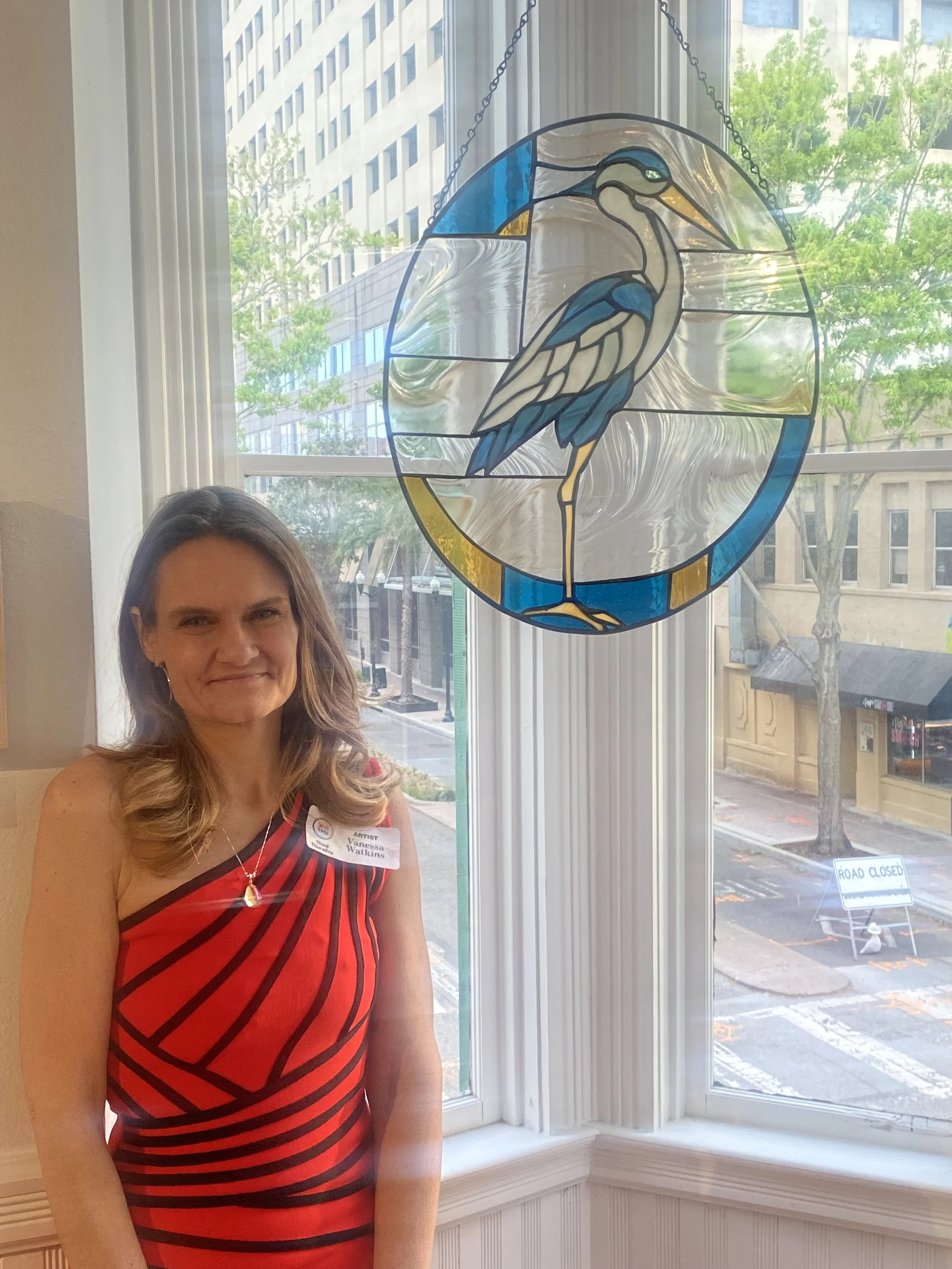 A woman with long, light brown hair smiling, wearing a red and black striped dress and a name tag that says Vanessa Watkins, stands inside near a large window with a stained glass pendant hanging above her. Outside the window, a city street is visible with a 'Road Closed' sign on the sidewalk, trees, and buildings across the street.