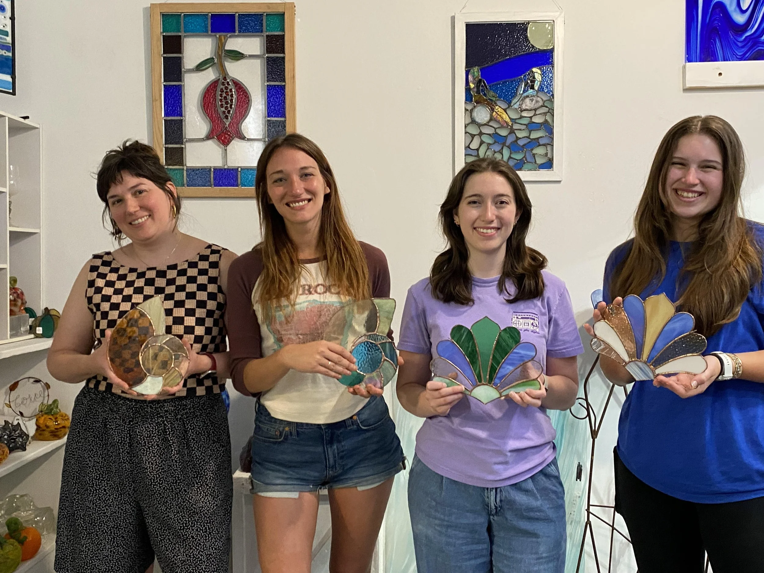 Four women standing in front of colorful stained glass art, holding glass butterfly craft projects.