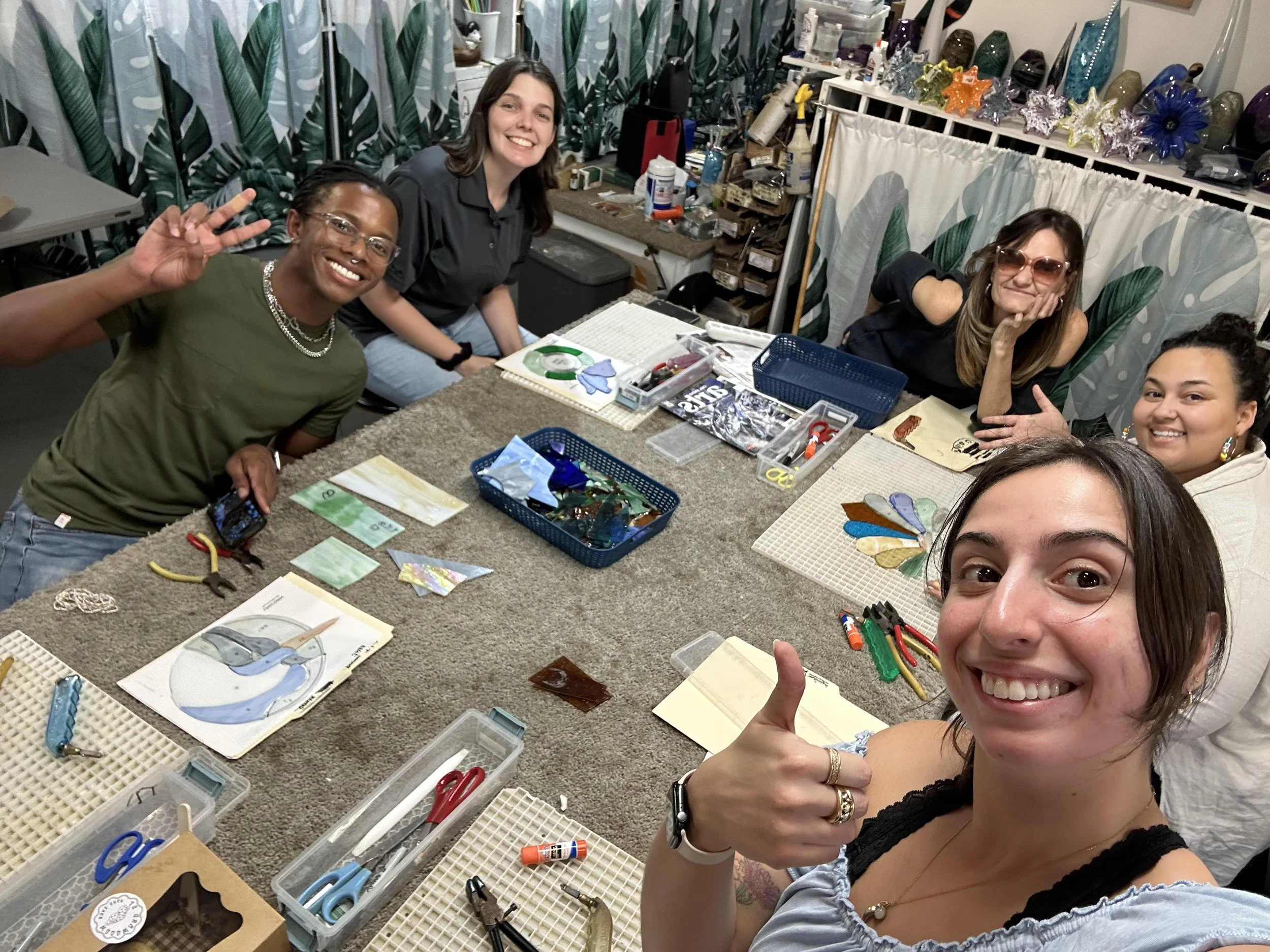 A group of four women sitting around a table with craft supplies, smiling and posing for a selfie. The table has various tools, papers, and decorative glass items.