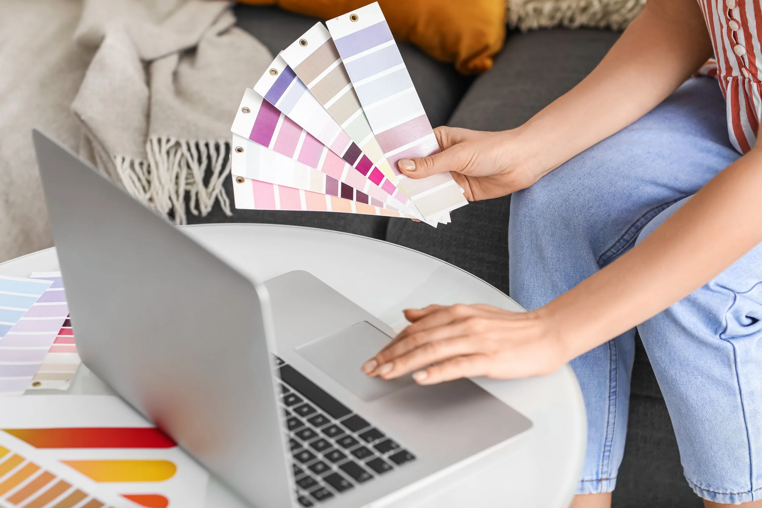 A person sitting on a couch arranging color swatches in front of a laptop on a coffee table.