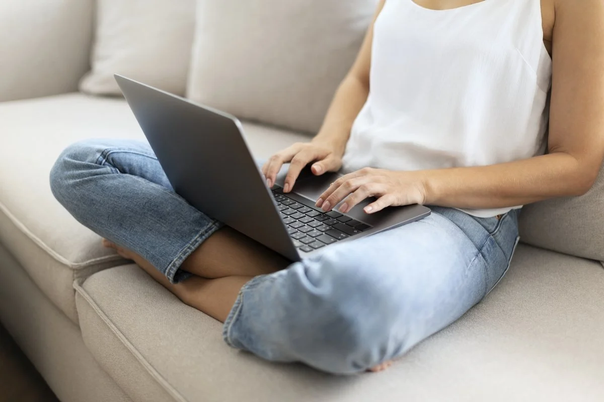 Person sitting cross-legged on a beige couch using a laptop computer.