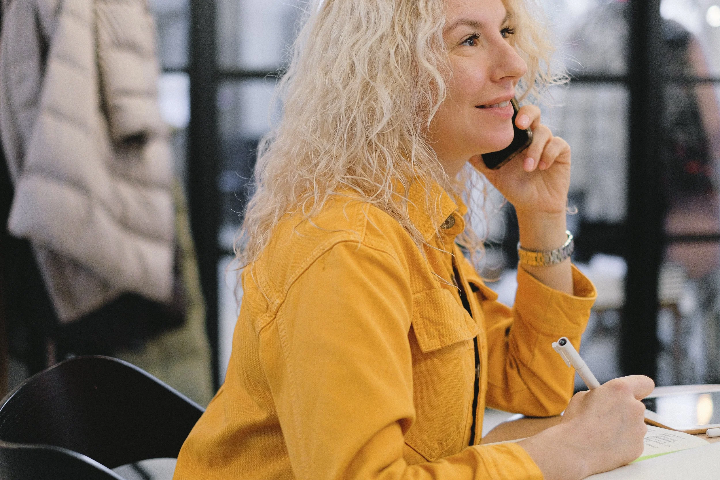 A woman with curly blond hair sitting at a table, talking on a cell phone, and taking notes with a pen.