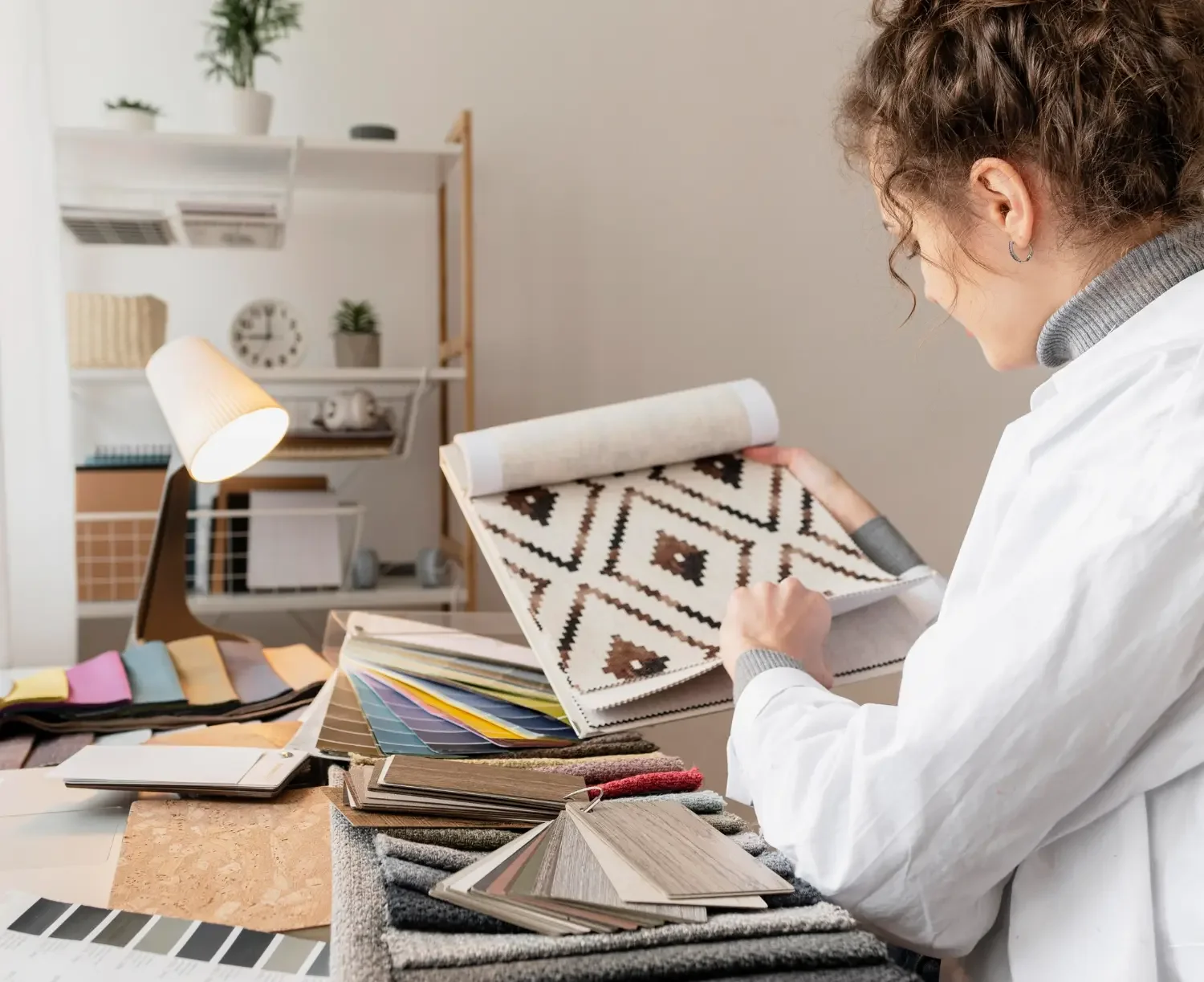 A woman in a white coat examining fabric or wallpaper samples at a workspace with color swatches, fabric samples, and design materials on a table, and a lit desk lamp in a modern, organized studio.