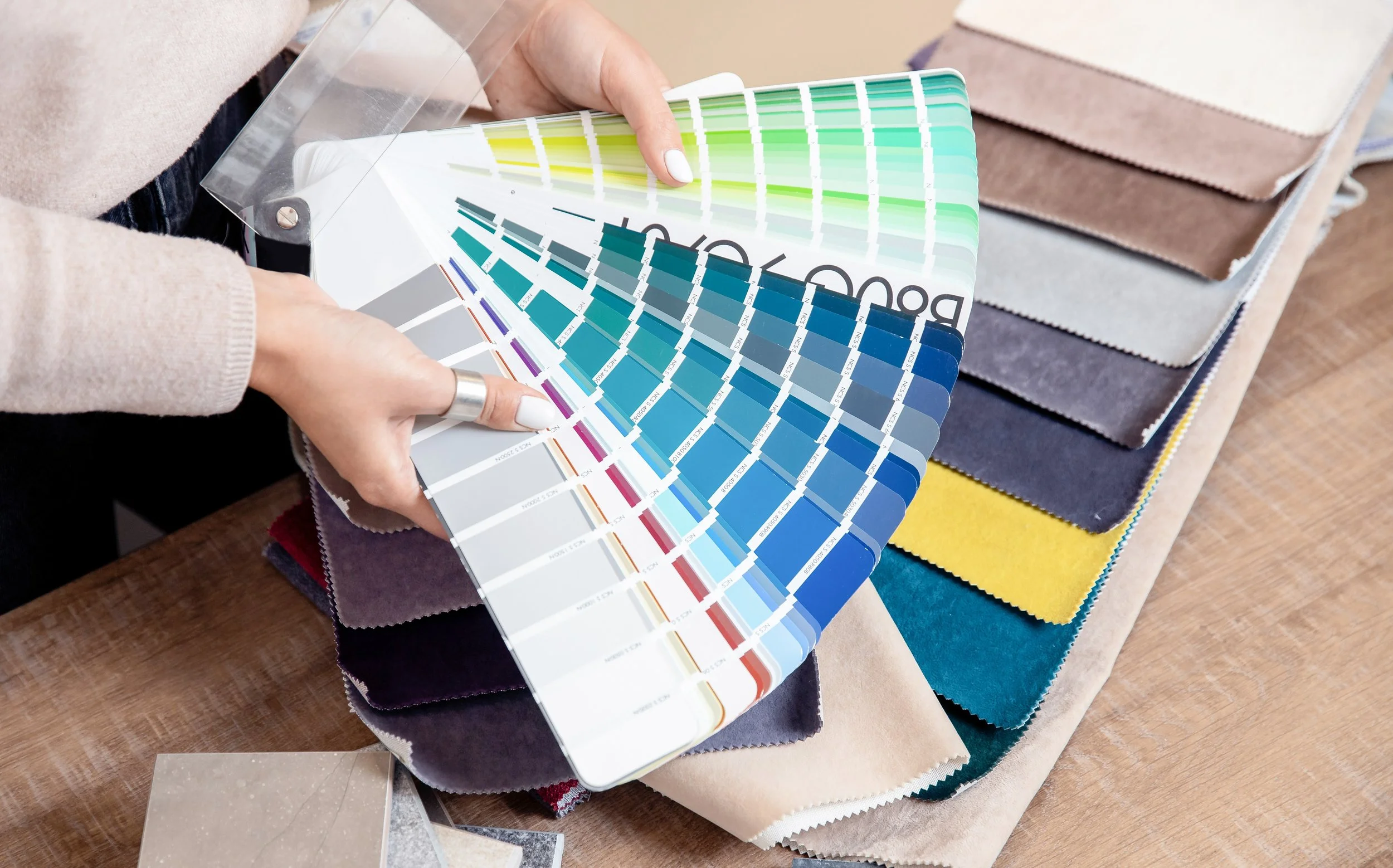 Person holding a color swatch fan with various fabric and material samples on a wooden table.