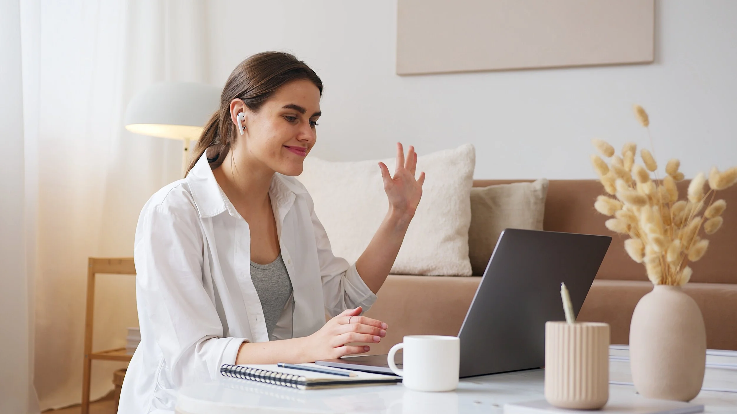 A woman sitting at a white desk on a video call, wearing wireless earbuds and waving, with a laptop, a mug, a notebook, and a vase with dried flowers in the foreground, in a cozy, well-lit living room.