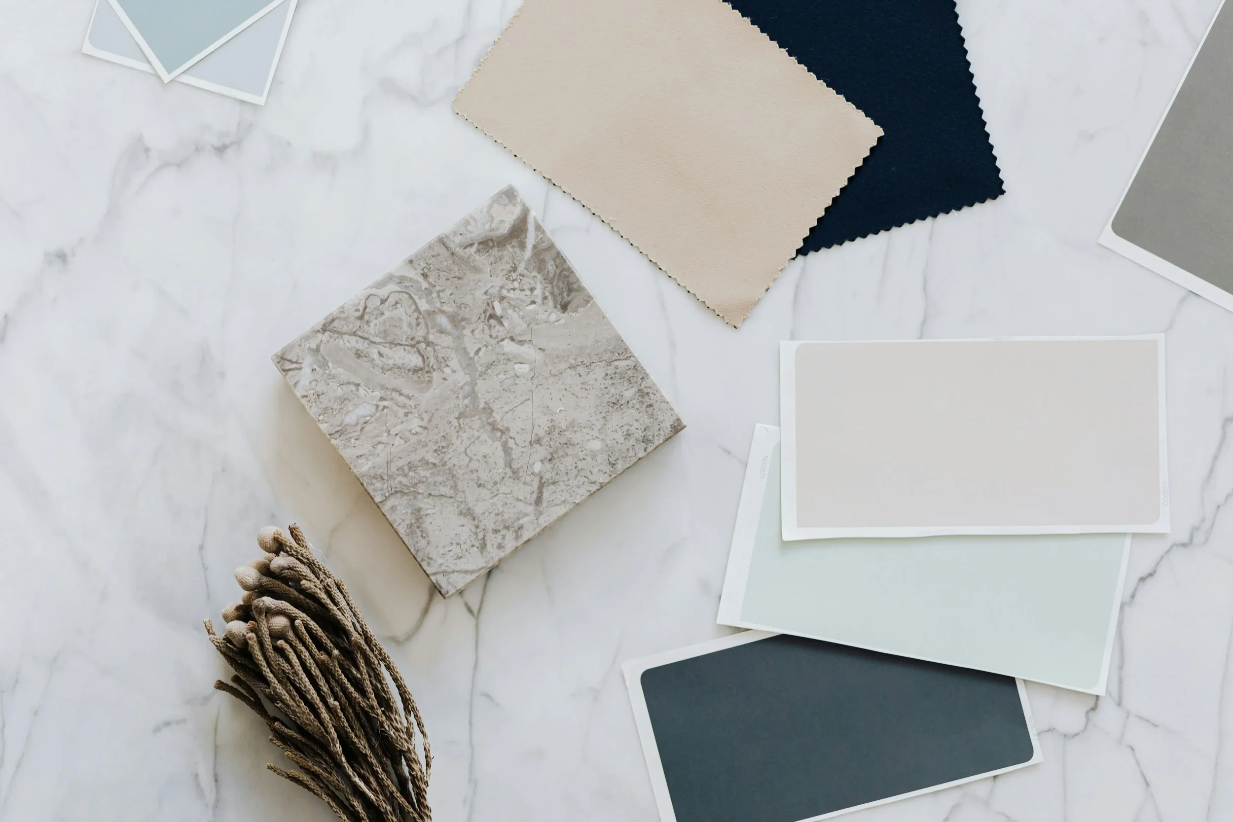 Samples of fabric, paint, and tile swatches on a white marble surface, with a bundle of dried flowers in the bottom left corner.