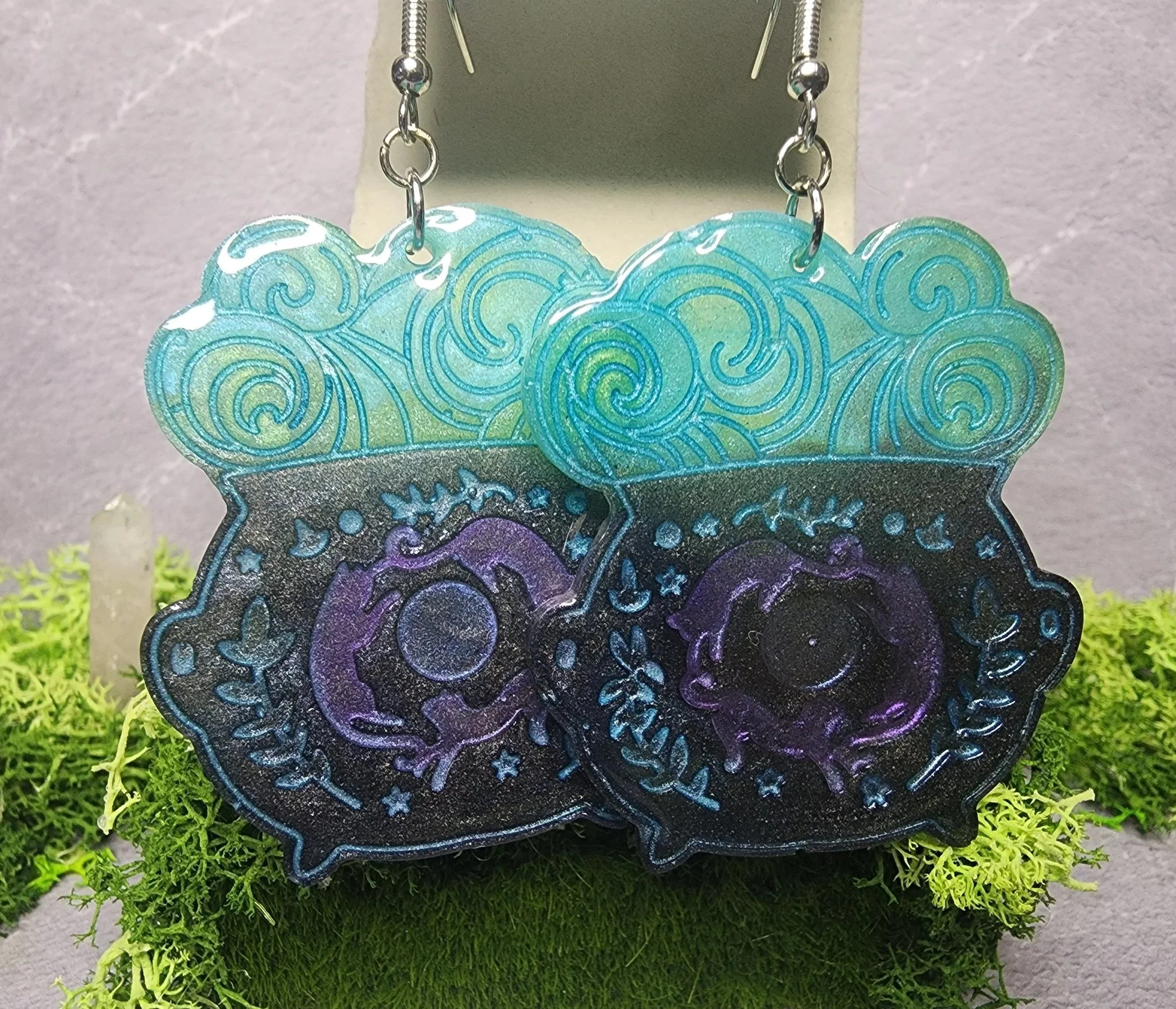 Handmade UV resin cauldron earrings with aqua-blue potion clouds, purple accents, and silver hypoallergenic hooks. up close hanging from display.
