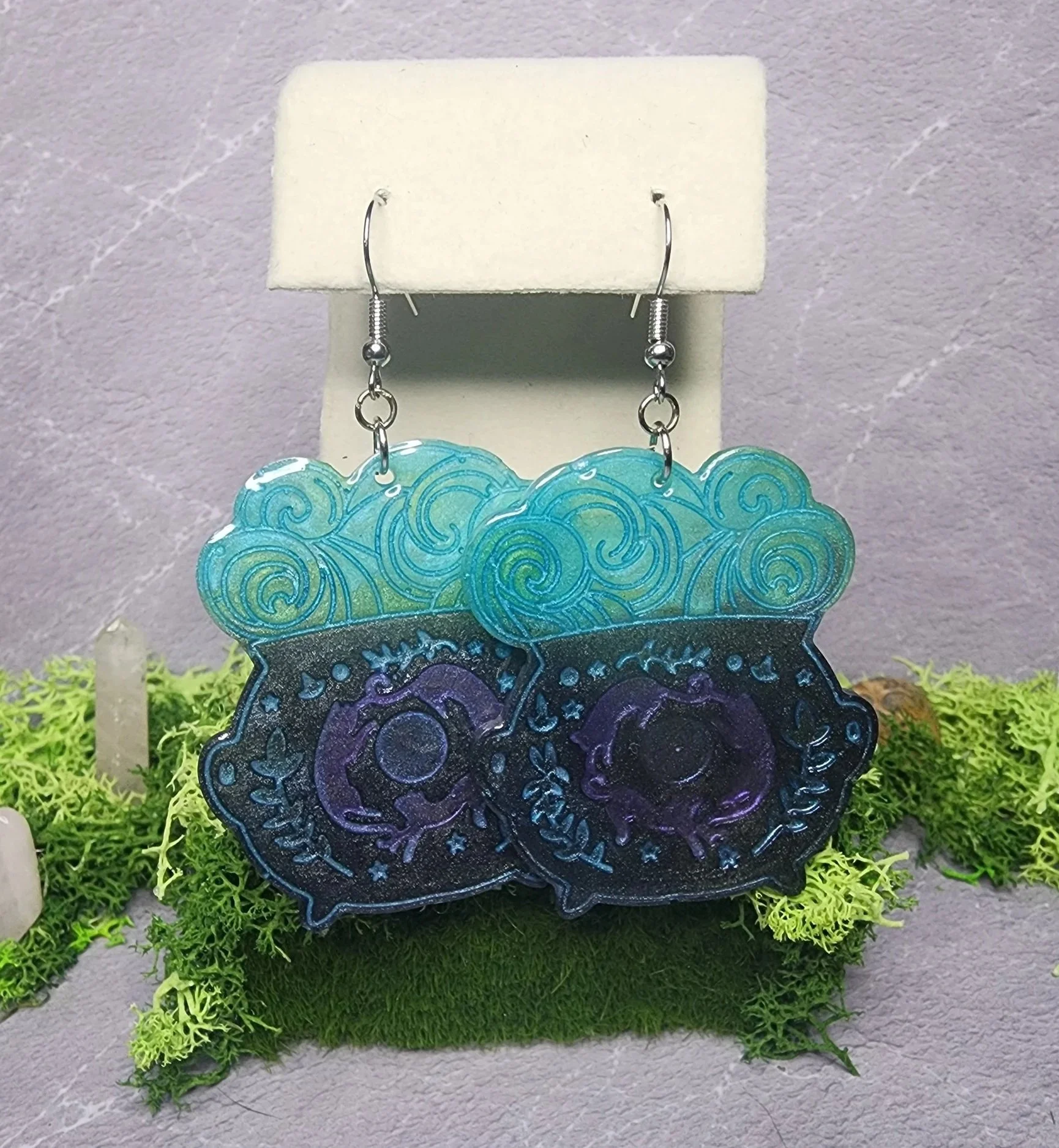 UV resin cauldron earrings with aqua-blue potion clouds, purple accents, and silver hypoallergenic hooks displayed hanging.