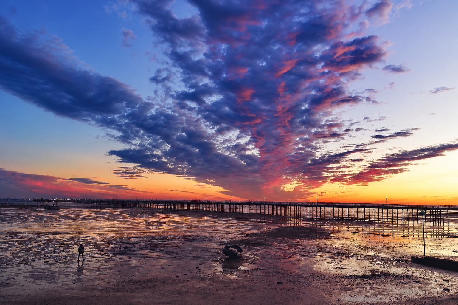 A dramatic sunset over the coastline, with vibrant purple, blue and orange clouds reflected across the wet sand at low tide. A solitary person walks along the exposed shoreline beside a small boat, while a long pier stretches towards the horizon.