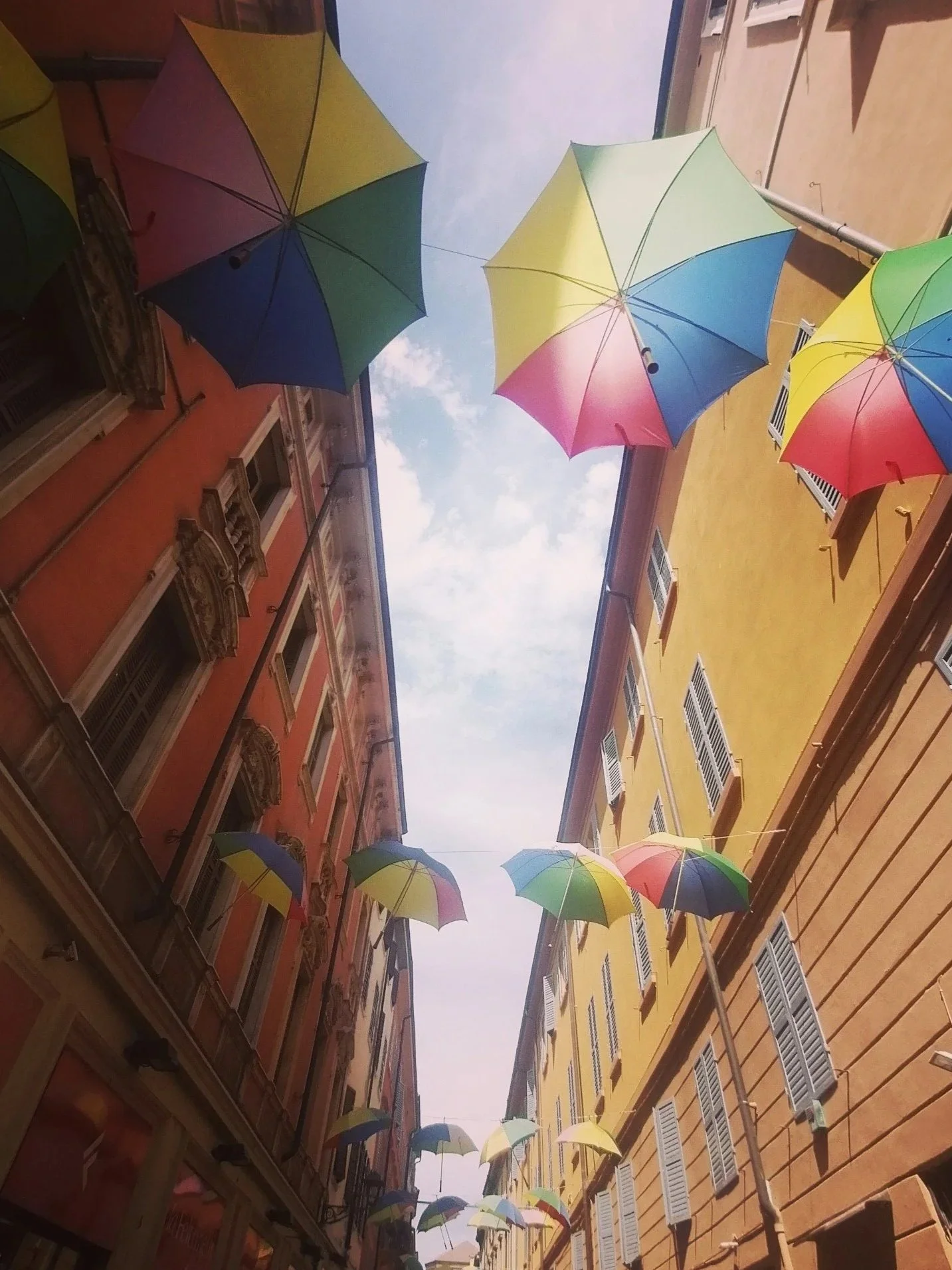 A narrow historic street with colourful umbrellas hanging overhead between pastel buildings, creating a vibrant canopy beneath the open sky.