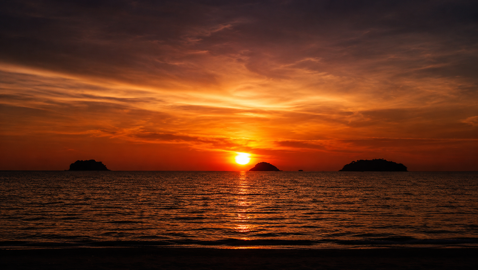 A warm sunset reflected across calm sea, with three distant islands beneath a glowing evening sky.