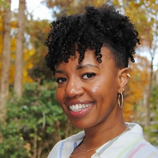 A woman with short, curly black hair smiling outdoors during fall, wearing hoop earrings and a white shirt.
