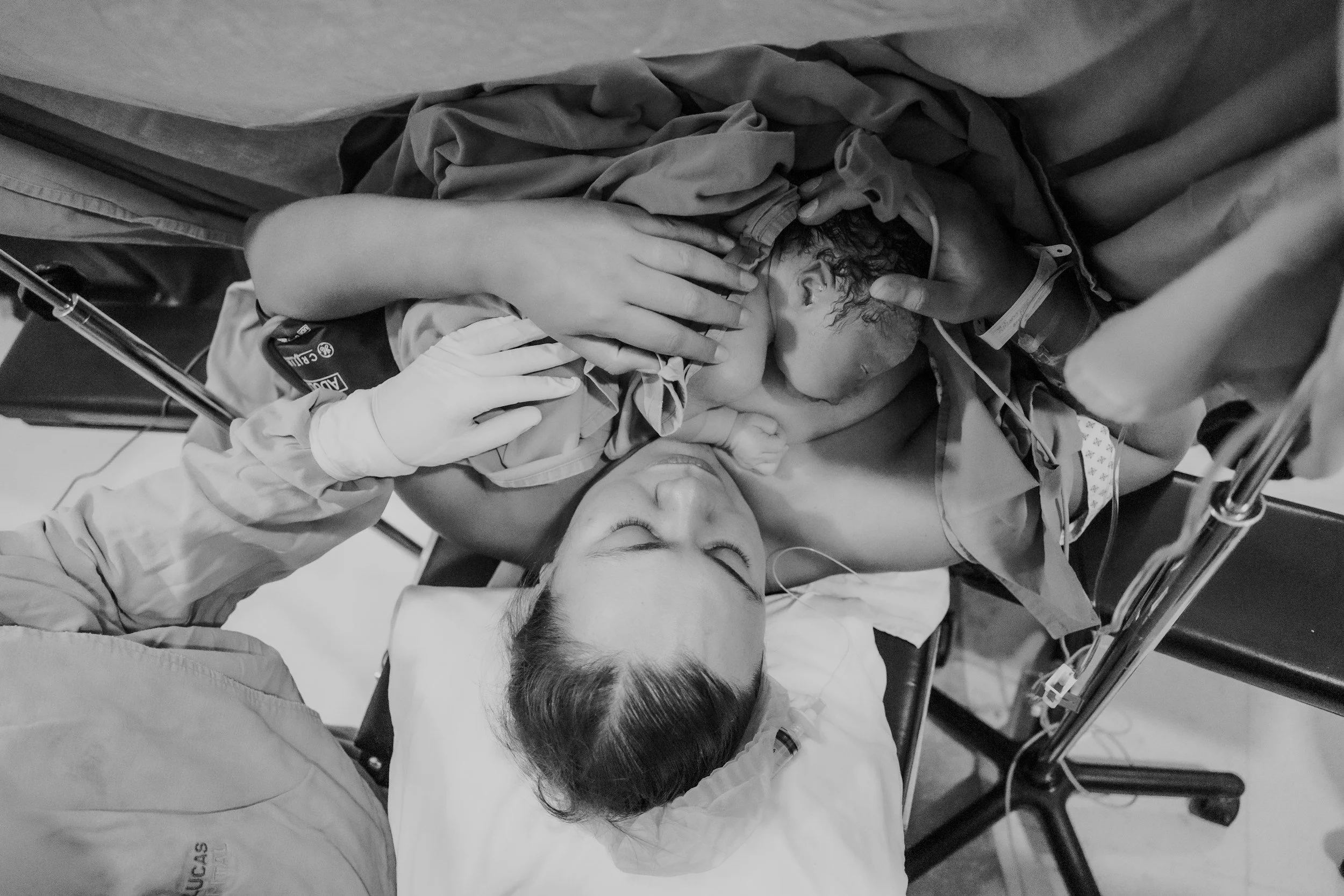 A woman lying on a hospital bed with her eyes closed, wearing a nasal oxygen tube. Several medical staff members are attending to her, one holding her head and another taking her hand. The scene appears to be in a medical or emergency setting.