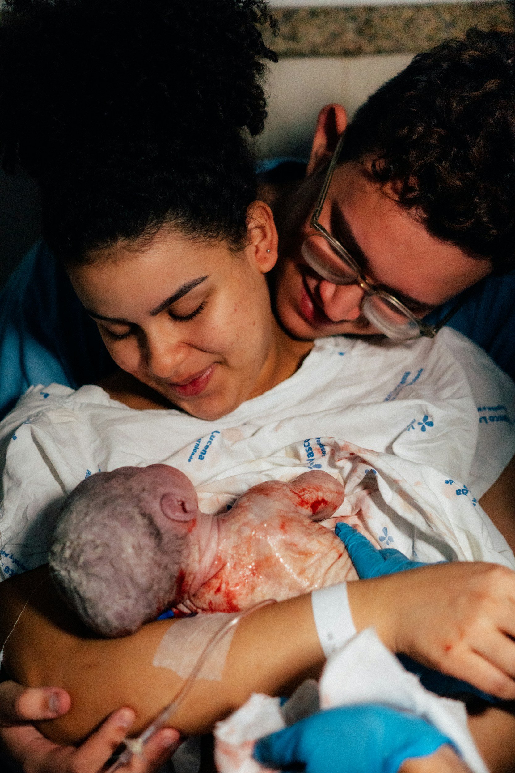 A couple in a hospital room holding a newborn baby, with the woman and the man smiling and looking at the baby, who is wrapped in a hospital blanket.