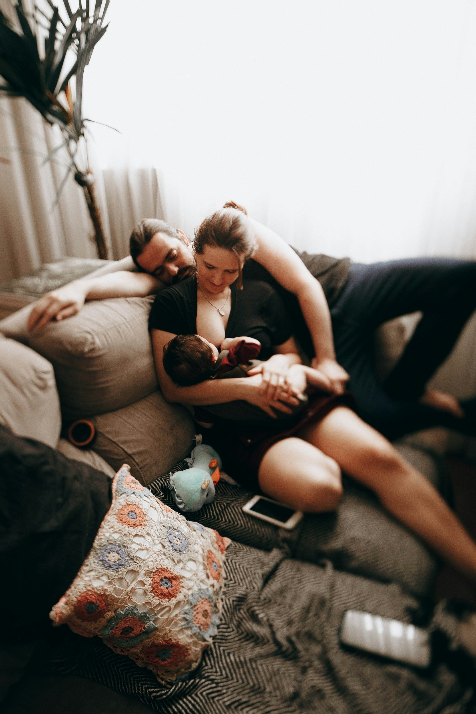 A man and a woman are lying on a couch, with the man resting his head on a pillow and the woman holding a baby. The woman is breastfeeding the baby, and there are everyday objects like a smartphone and a blanket nearby.