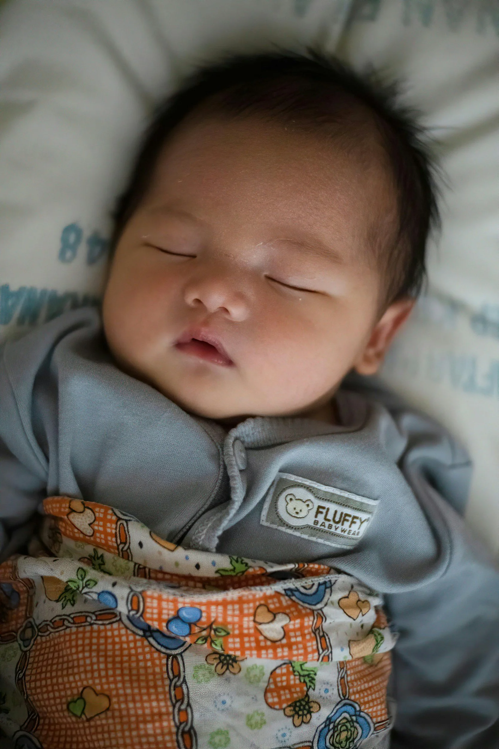 A sleeping baby with closed eyes, wearing a gray jacket with a "Fluffy Babywear" label, lying on a blanket with colorful cartoon designs.