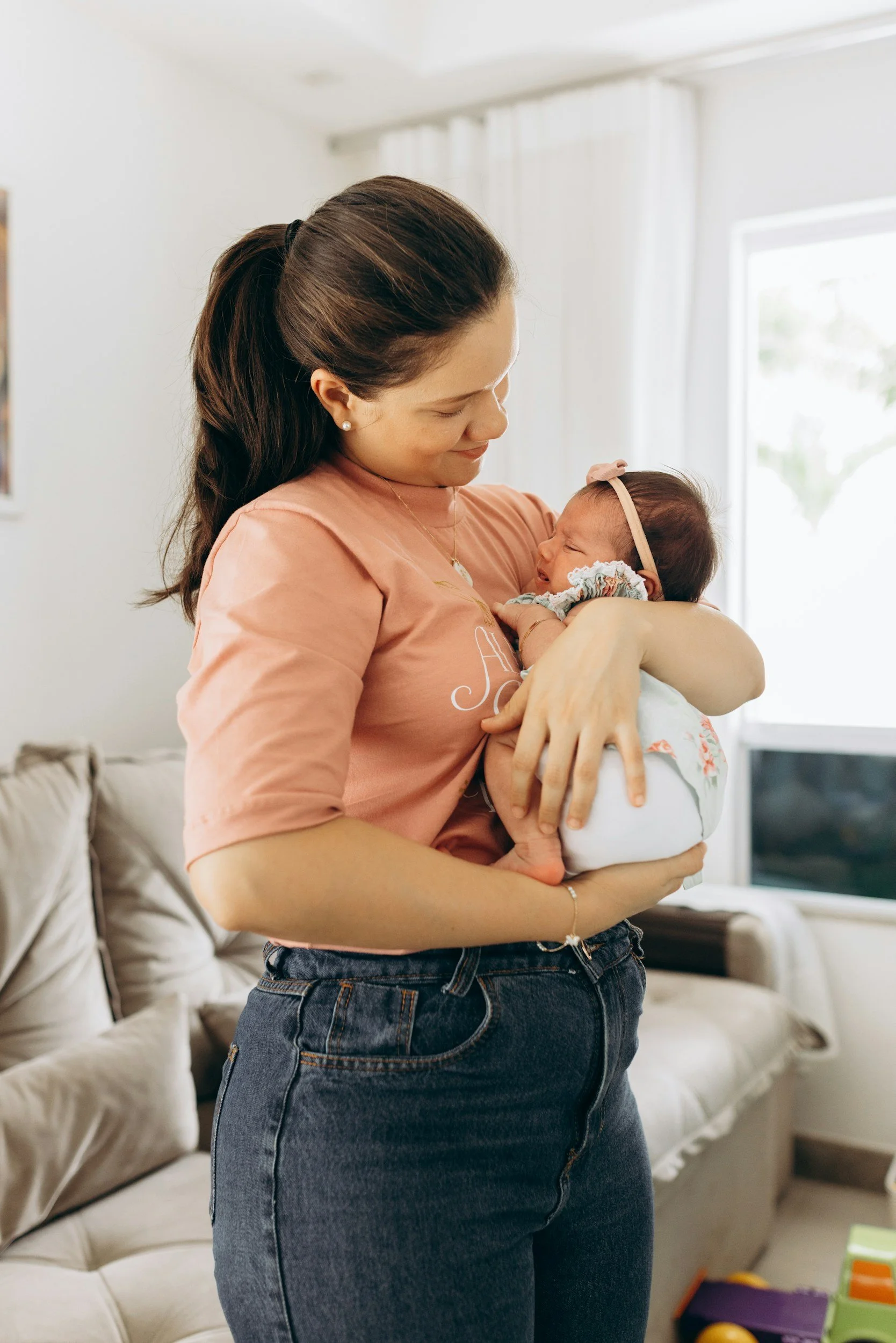 A woman with dark brown hair in a ponytail holding a newborn baby girl. The woman is smiling while looking at the baby, who appears to be crying. The background shows a living room with a beige couch, a window with white curtains, and some colorful toys on the floor.