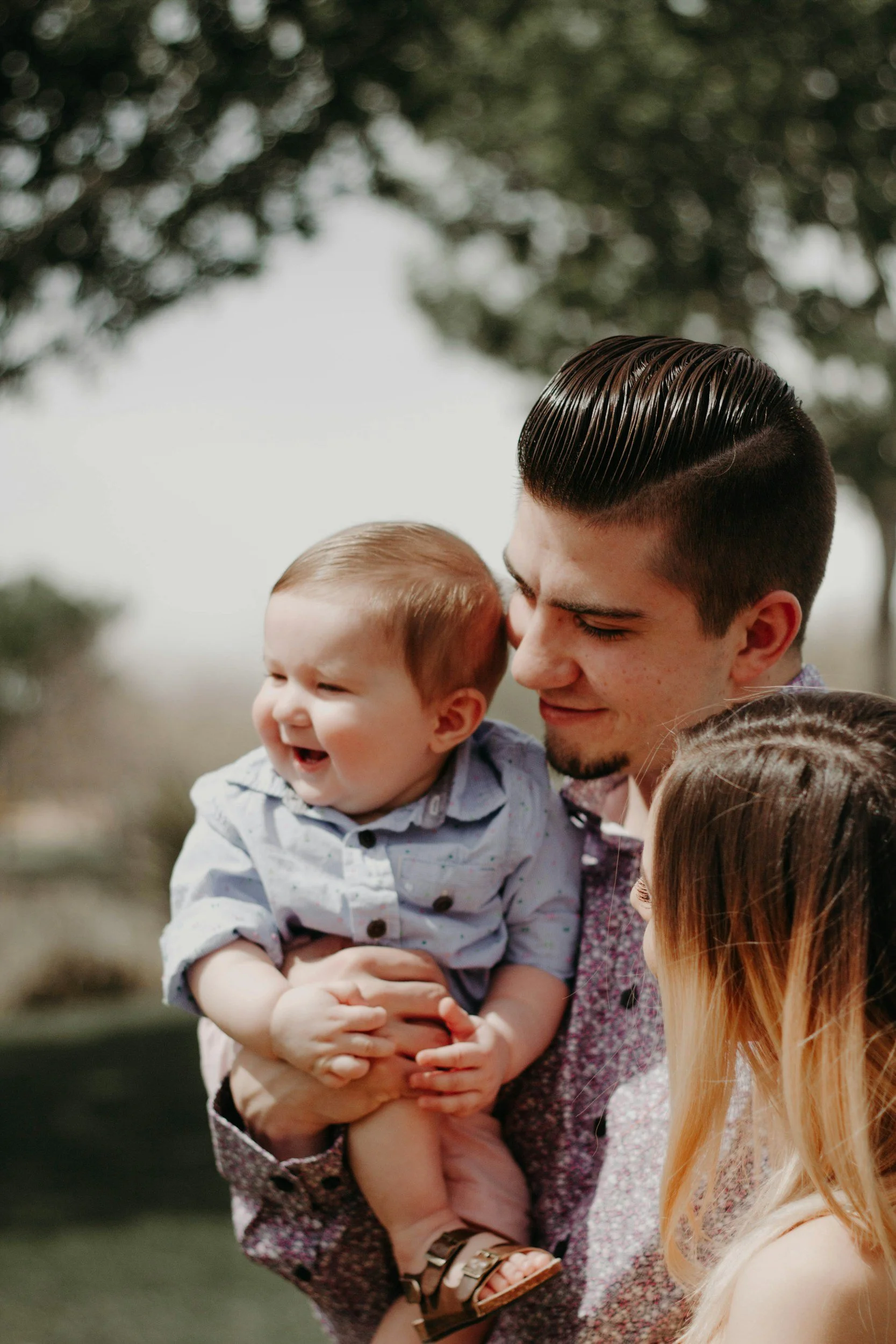 A man holding a smiling toddler outdoors, with a woman and blonde girl nearby, all smiling and enjoying a sunny day.