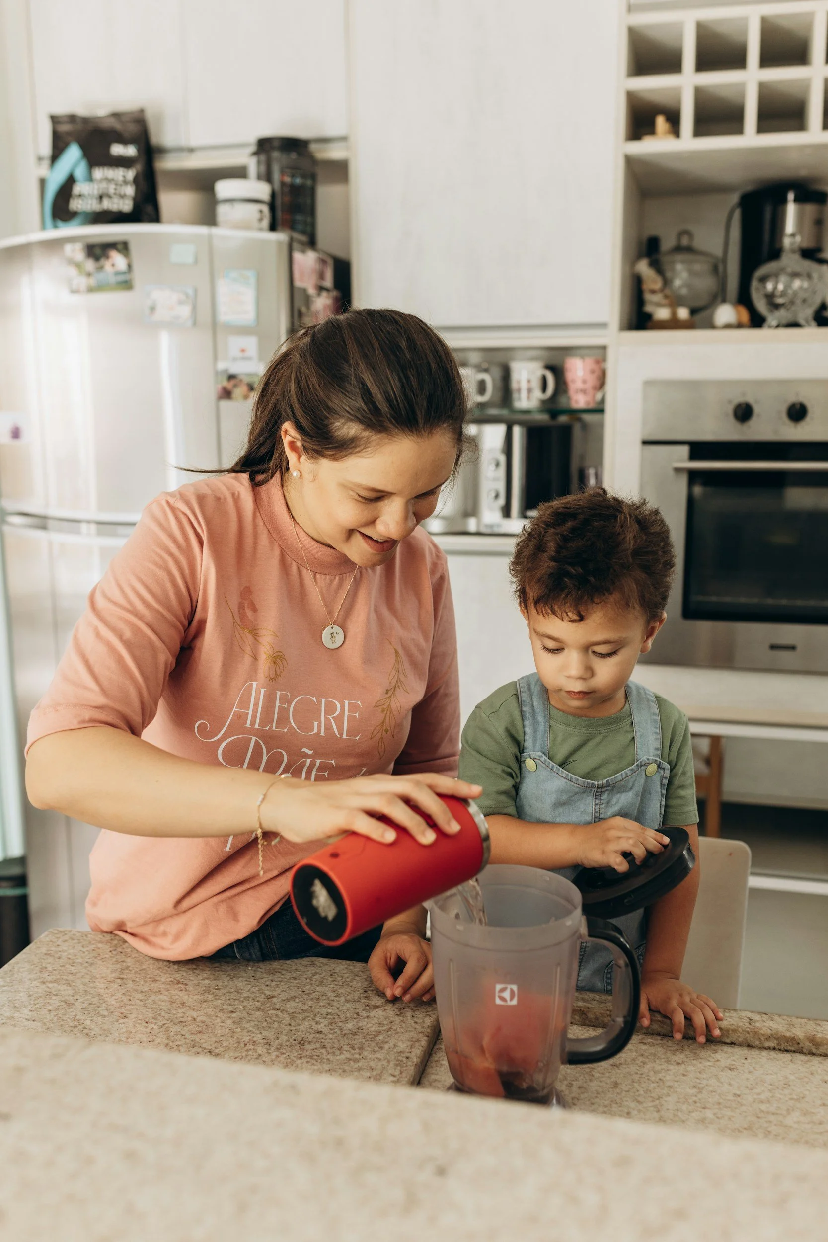 A woman and a young boy in a kitchen, pouring a liquid into a blender while the boy looks on.