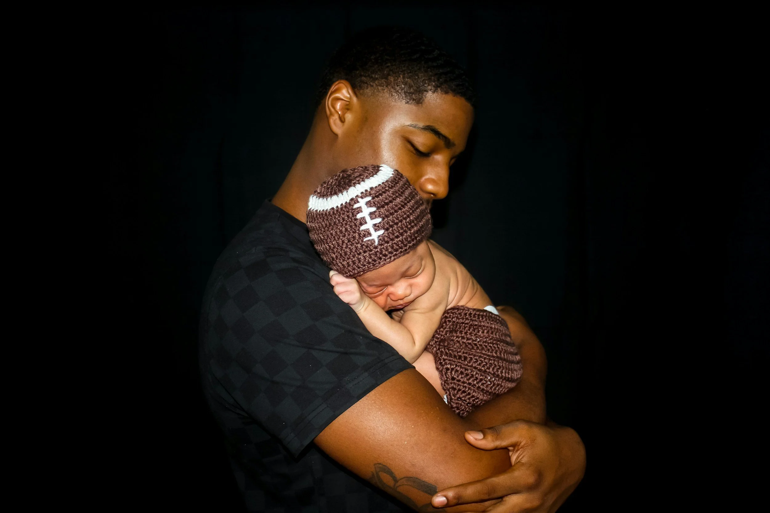 A young man with short curly hair and dark skin is holding a sleeping baby with light skin, dressed in a brown knitted hat and sweater, against a black background. The man has his eyes closed and is gently embracing the baby.