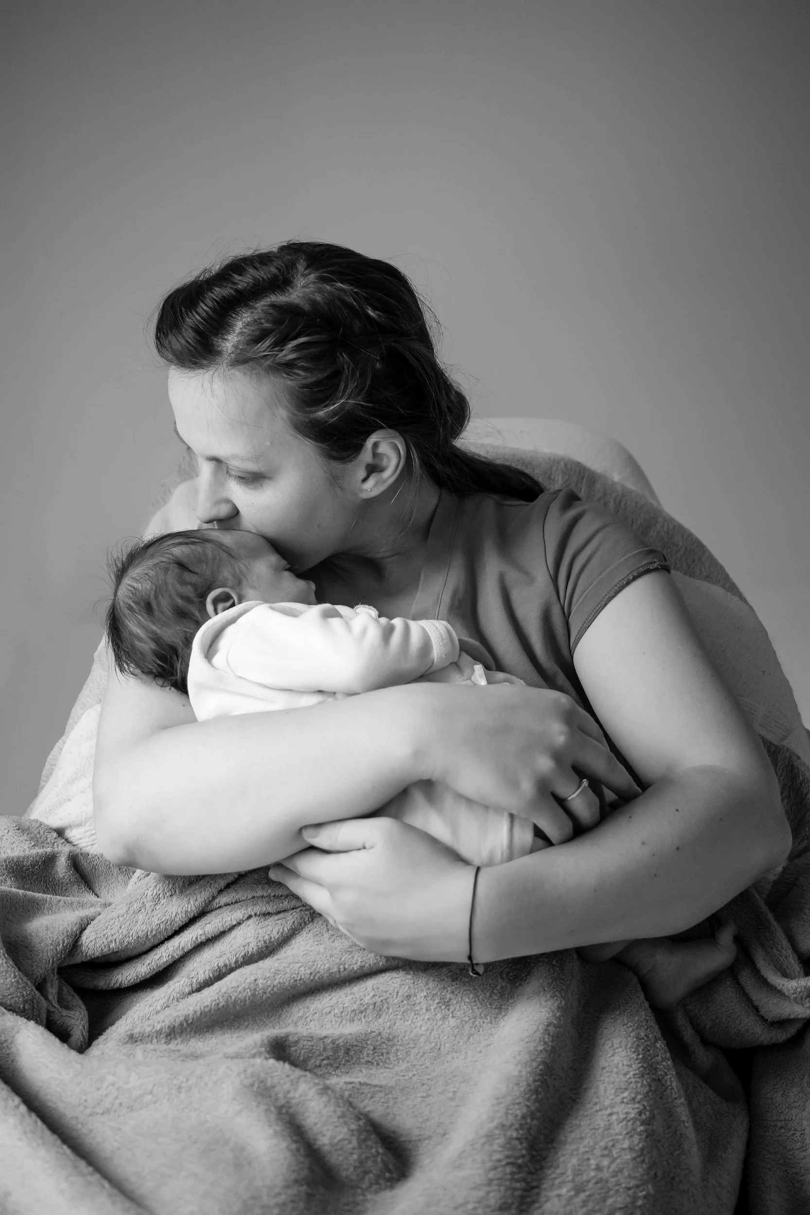 A woman with dark hair styled in a vintage wave, holding and kissing a sleeping baby in her arms. The scene is in black and white, and the woman wears a short-sleeved top, sitting on a lap with a blanket draped over her lap.