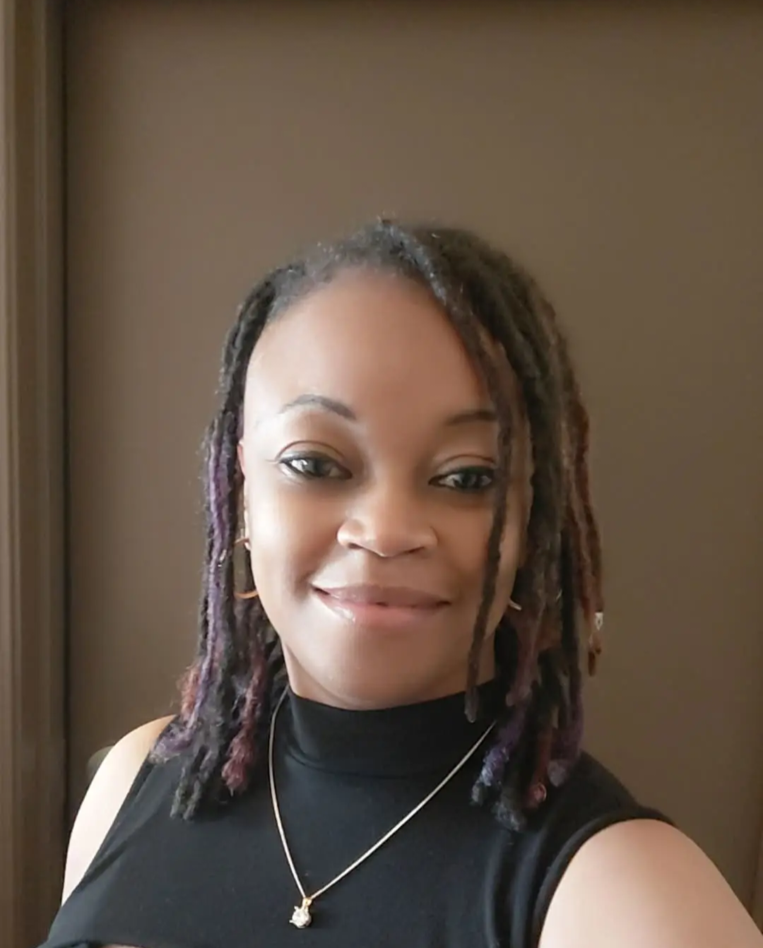 A woman with medium-length dreadlocks, some with purple highlights, wearing a black sleeveless top, gold hoop earrings, and a delicate gold necklace with a pendant, smiling at the camera.