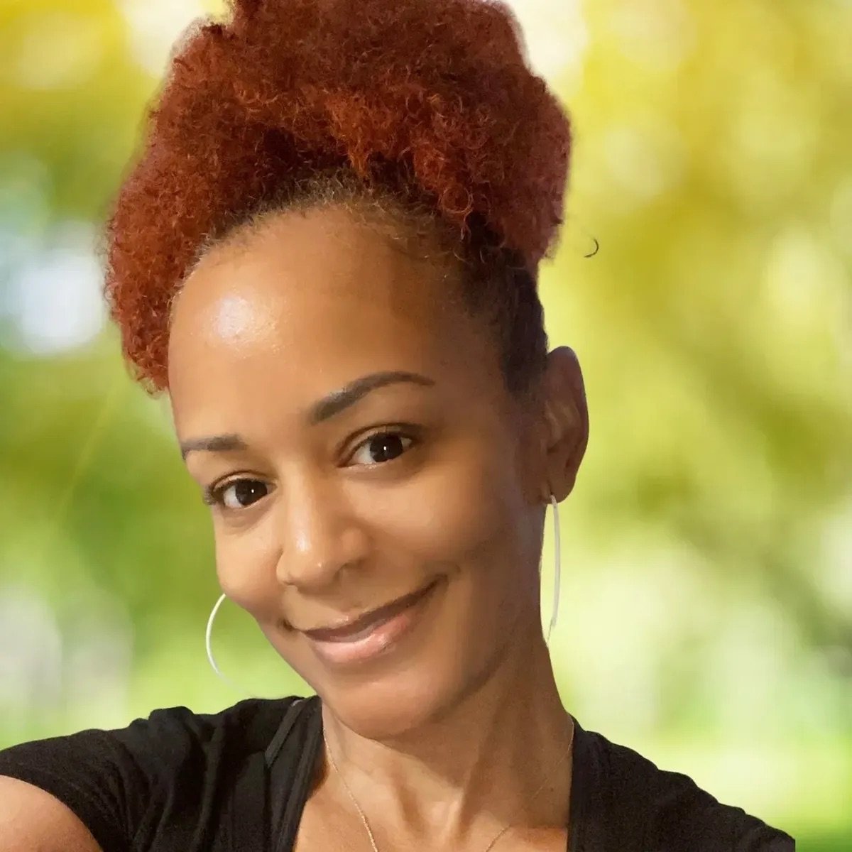 A woman with dark skin and reddish-brown curly hair styled in an updo, smiling softly at the camera. She is wearing hoop earrings and a black top against a blurred green and yellow background.