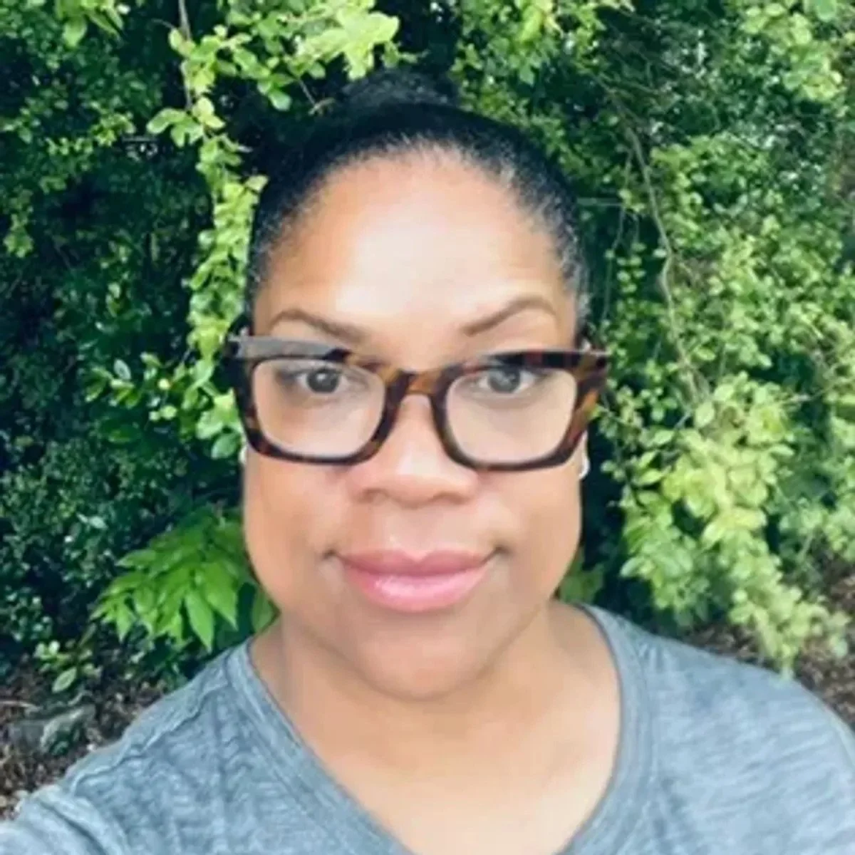 A woman with glasses smiling outdoors with green foliage in the background.
