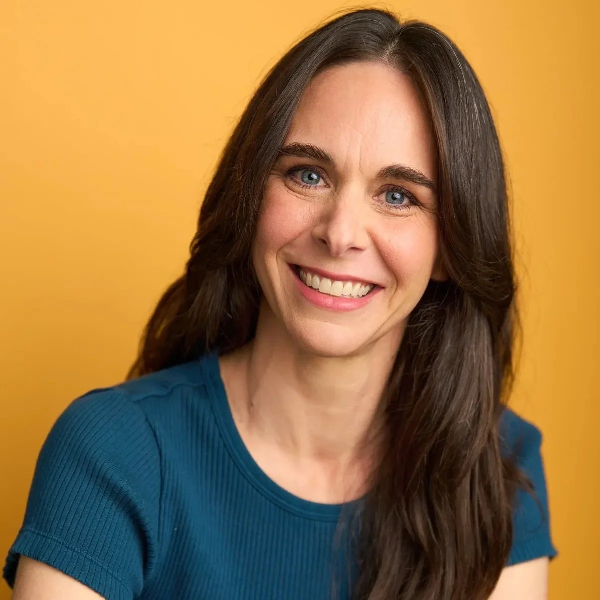 A woman with long brown hair and blue eyes smiling at the camera against a yellow background.
