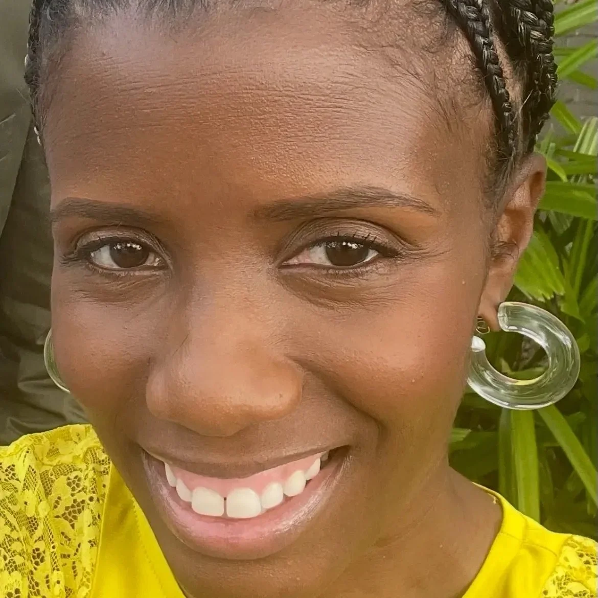 Close-up of a smiling woman with dark skin, braided hair, wearing large clear hoop earrings and a yellow lace shirt, with green plants in the background.