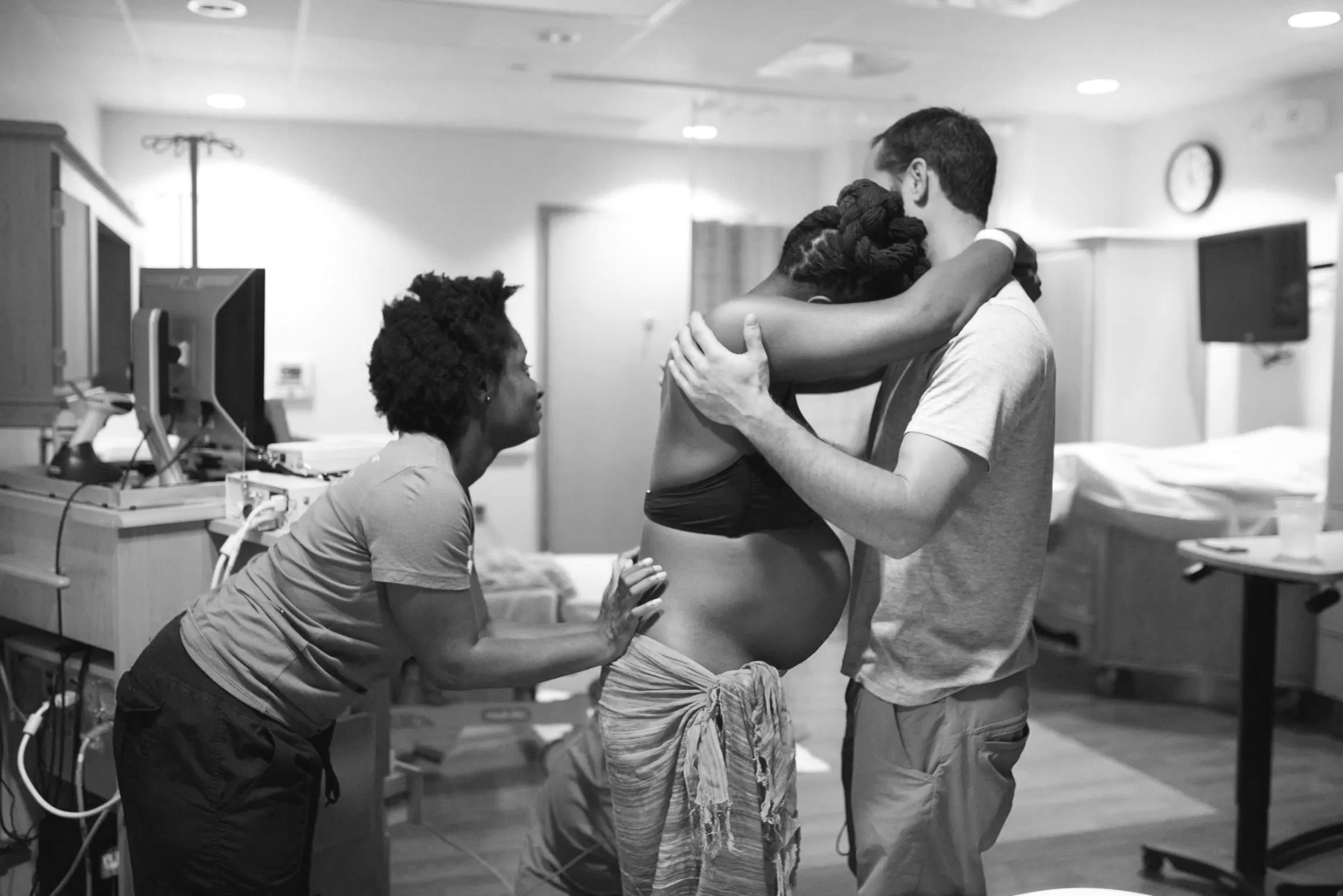 A black and white photo of a woman seated next to a hospital bed, holding hands with a woman in a hospital gown, while a man embraces a woman with braided hair, all in a hospital setting.