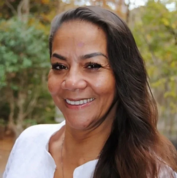 A woman with long dark hair smiling outdoors with trees and fall foliage in the background.