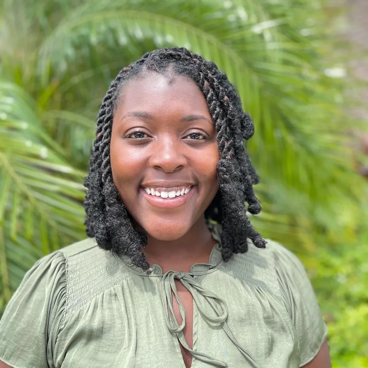 A smiling woman with dark curly hair styled in twists, wearing a light green blouse, standing outdoors with green tropical plants in the background.