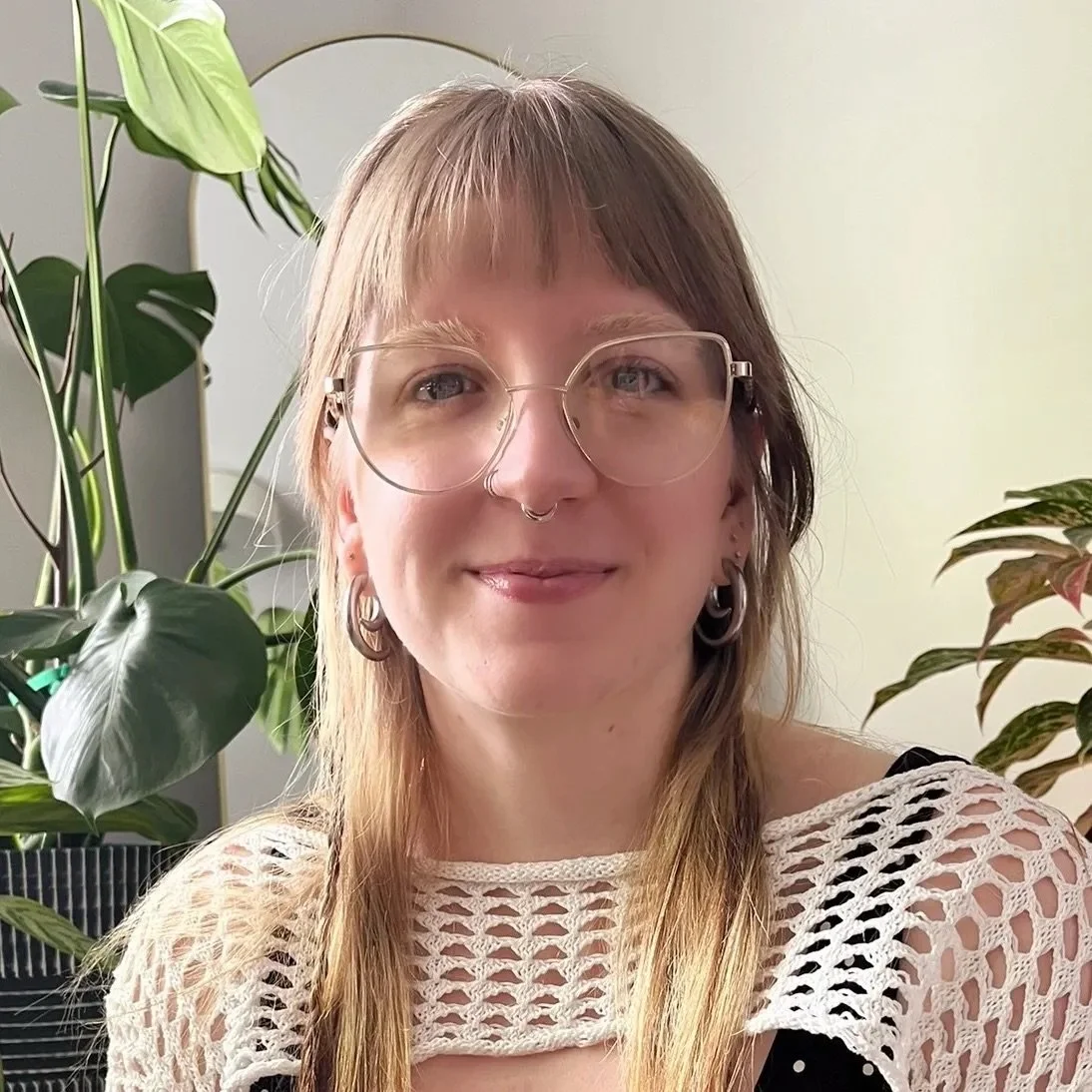 A woman with shoulder-length blonde hair, glasses, and hoop earrings sitting in front of green houseplants, wearing a crochet top.