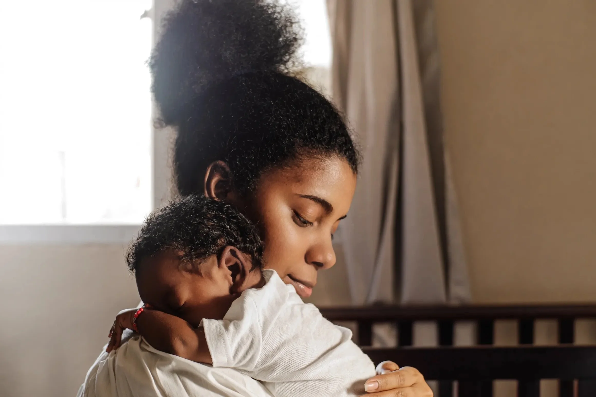 A woman with curly hair holding a young girl with curly hair in her arms as they both smile and look down, sitting indoors near a window and a bed.