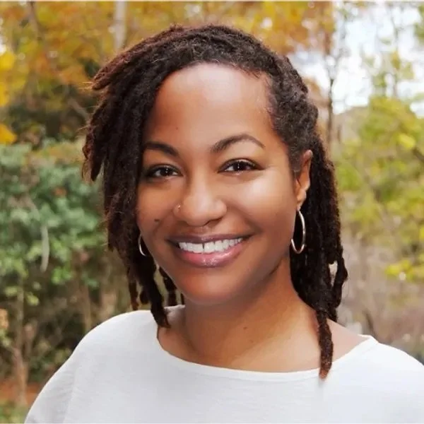 A woman with shoulder-length dreadlocks, hoop earrings, and a white top, smiling outdoors with trees in the background.