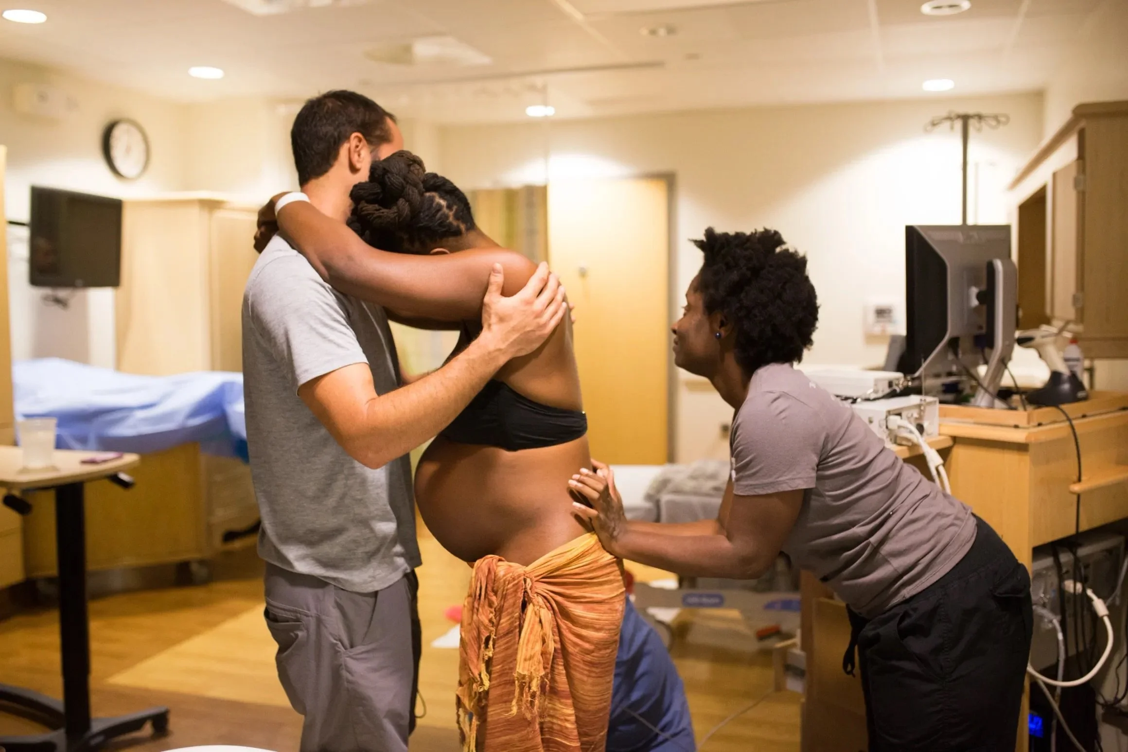 Woman with rhino horn-shaped hair hairstyle, pregnant, being examined by a healthcare professional in a medical setting, with two other women assisting.