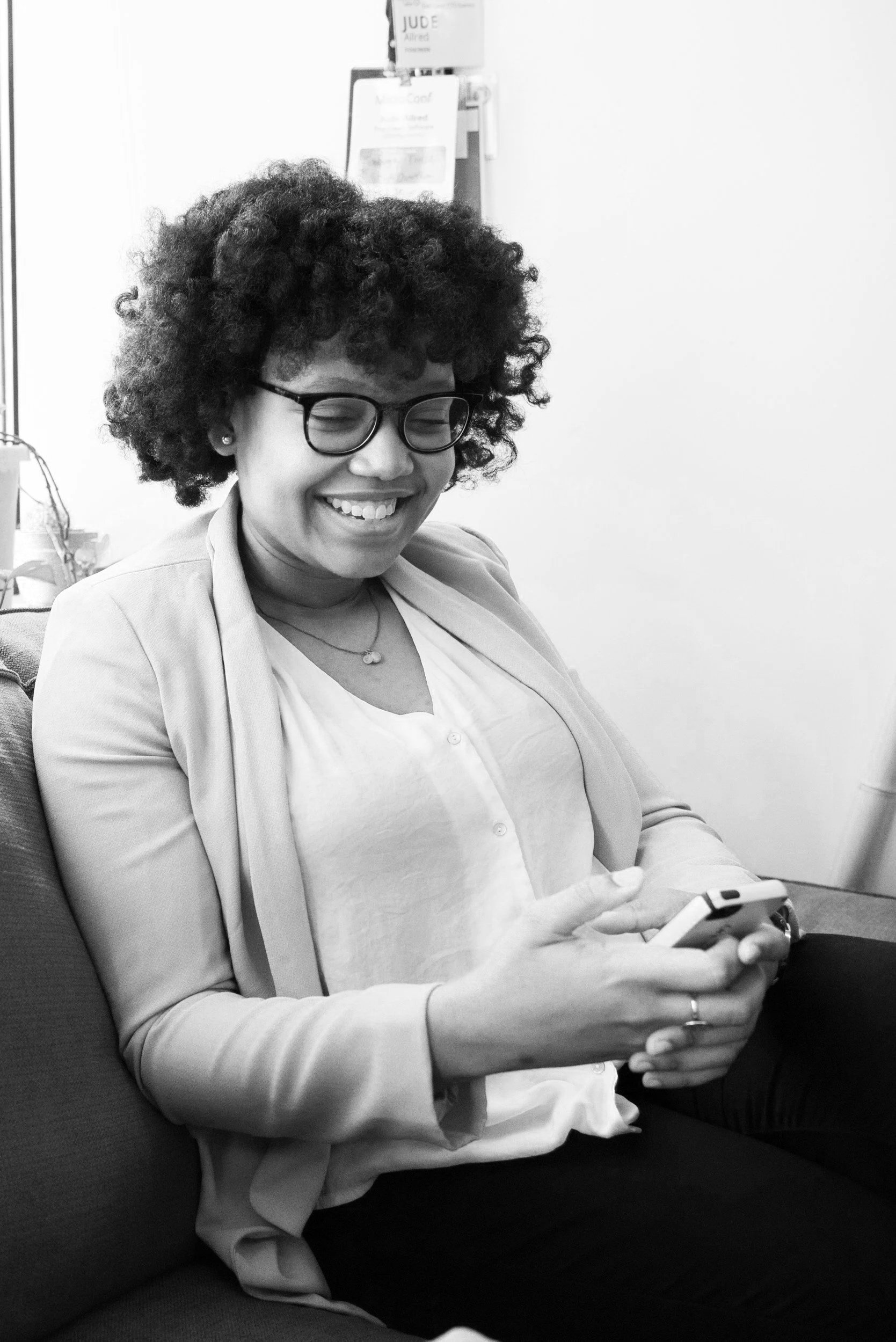 A woman with curly hair, glasses, and earrings smiling while looking at her phone. She is sitting on a chair with a plain wall behind her.
