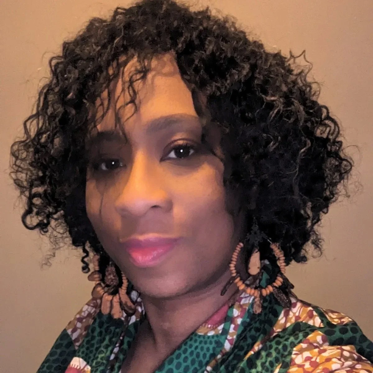 A woman with dark, curly, shoulder-length hair, wearing large wooden hoop earrings and a colorful patterned top.
