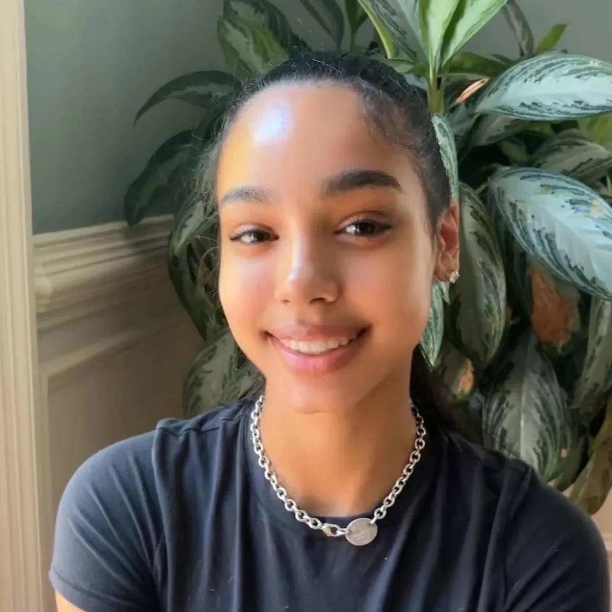 A young woman with dark hair tied back, smiling, wearing a black t-shirt, a silver chain necklace, and earrings, posing in front of a large green leafy plant indoors.