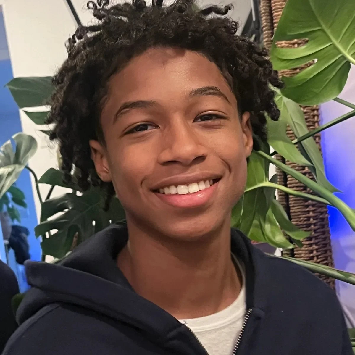 A smiling young man with curly hair standing in front of green tropical plants indoors.