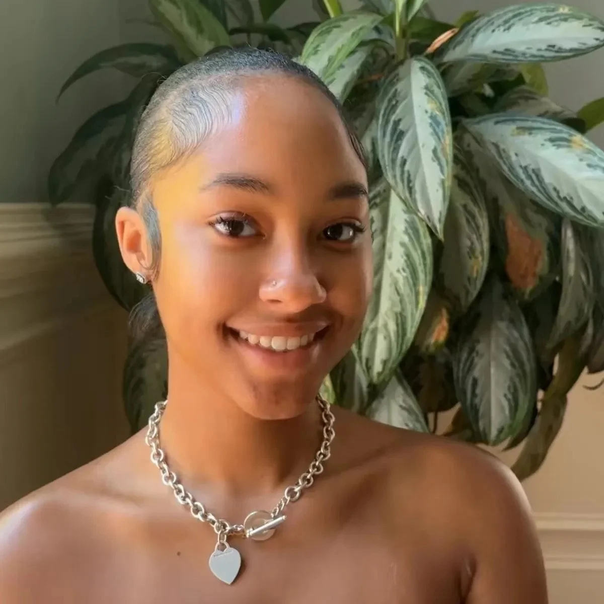 A young woman with a short, slicked-back hairstyle, smiling, wearing a chunky silver chain necklace with a heart pendant, standing indoors in front of a large green houseplant.