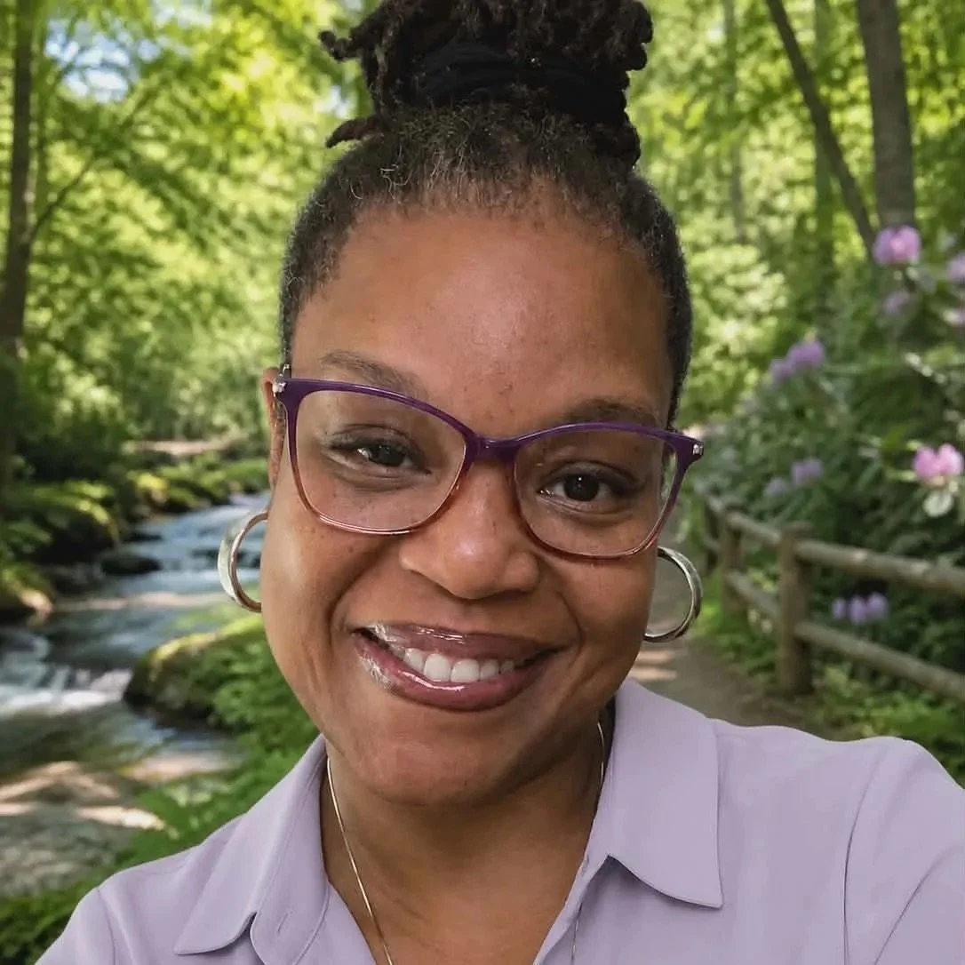 A woman smiling outdoors in front of a forest scene with a stream and purple flowers.