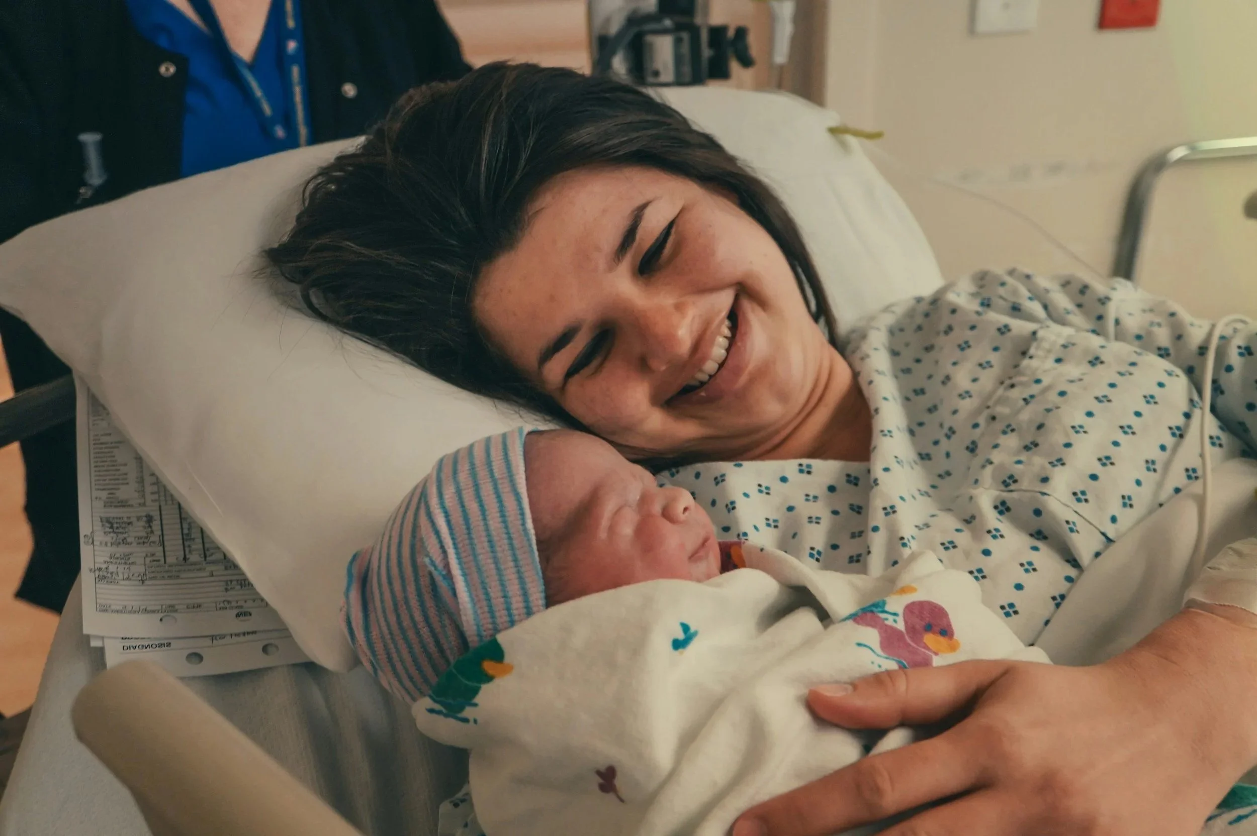 A woman smiling in a hospital bed holding a newborn baby, with a person in a blue shirt and dark jacket at the bedside.