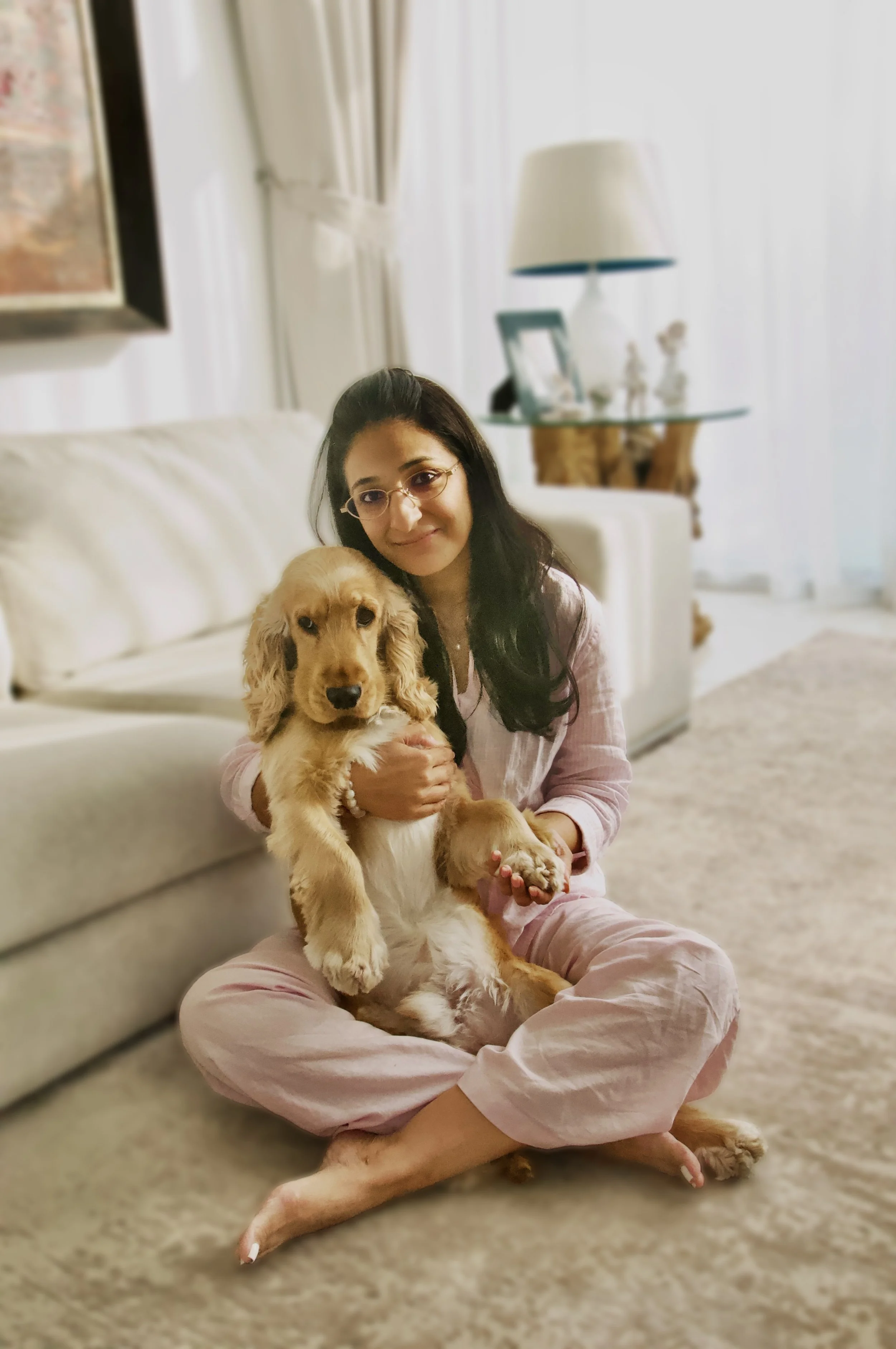 A woman with long dark hair and glasses sitting on a beige carpet, holding a golden retriever puppy. She is wearing pink pajamas, and has her legs crossed. The background shows a white sofa, a glass side table with a lamp, picture frames, and decorative items in a living room setting.