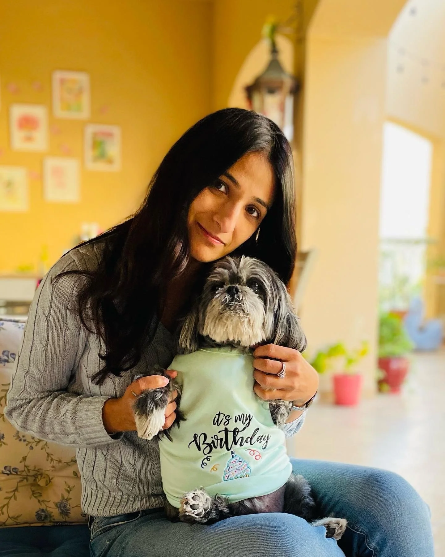 A woman sitting on a couch holding a small dog wearing a light green birthday shirt with birthday-themed text and logo, and a small heart-shaped ring on her finger, in a room with yellow walls and colorful decorations.