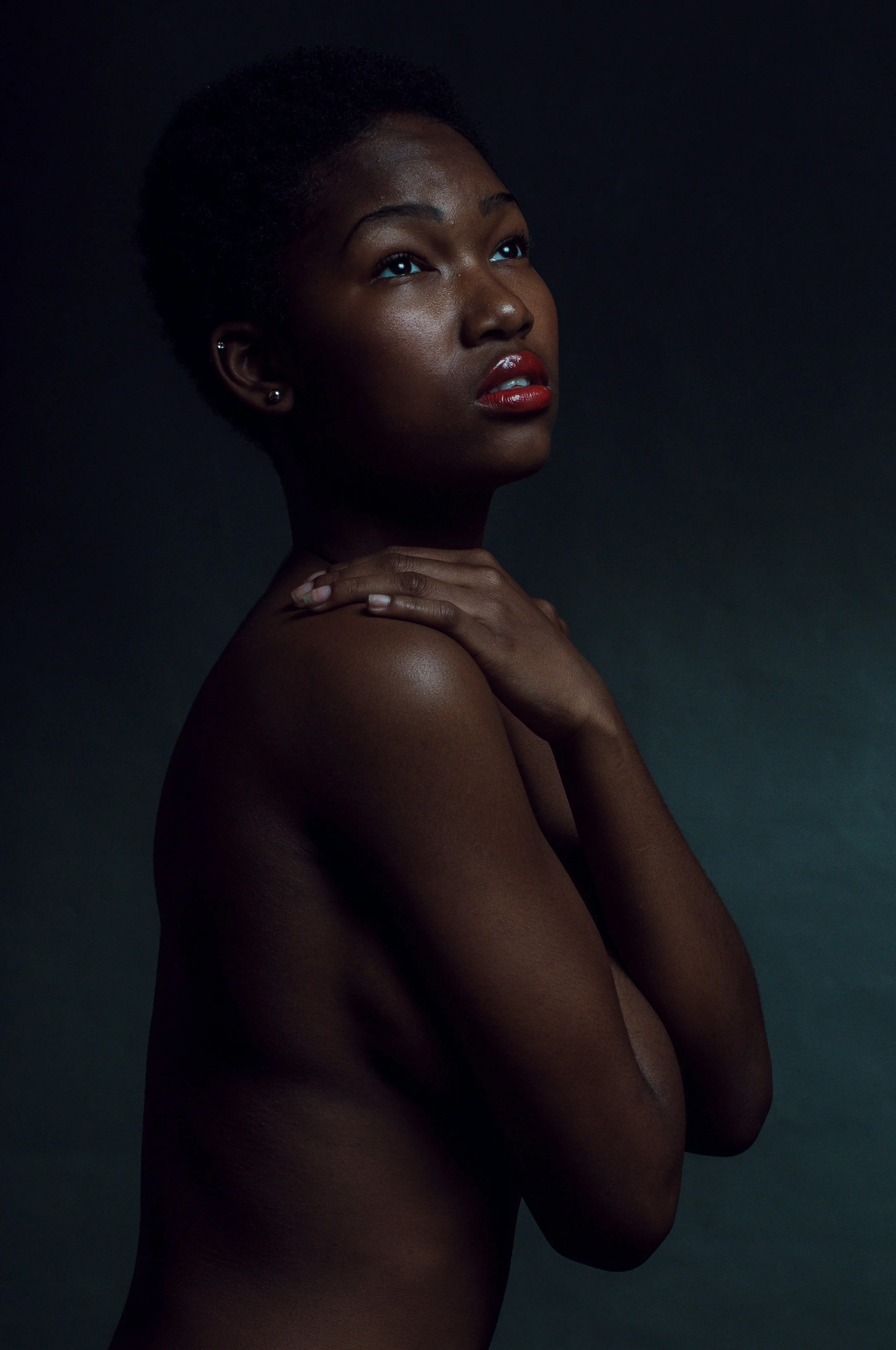 A portrait of a woman with dark skin, short curly hair, and red lipstick, looking upwards with her hands gently resting on her shoulder, set against a dark background.