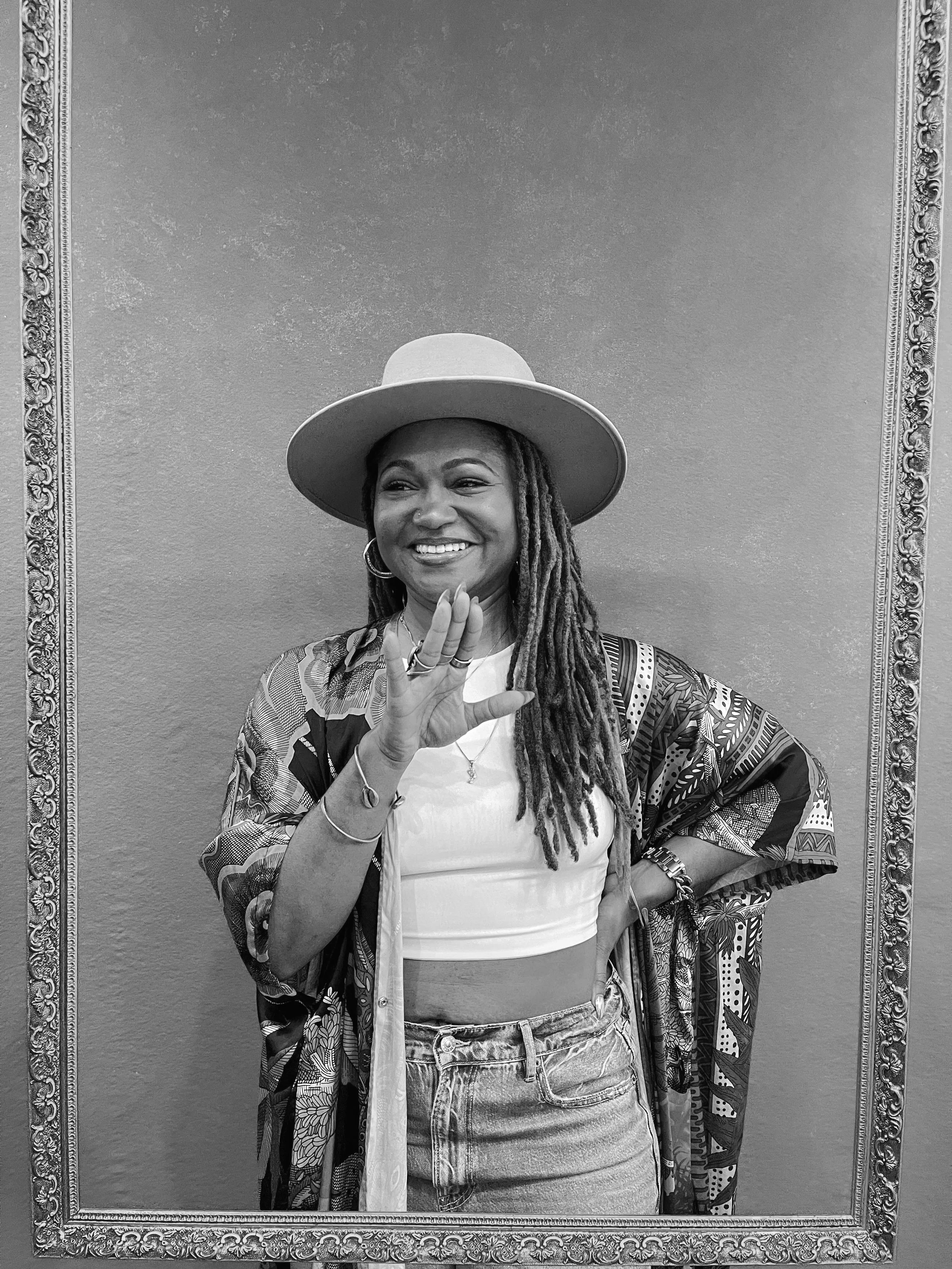 A woman with dreadlocks smiling, wearing a wide-brimmed hat, hoop earrings, a patterned kimono, a white crop top, and high-waisted denim shorts, standing against a plain wall with an ornate picture frame around her.